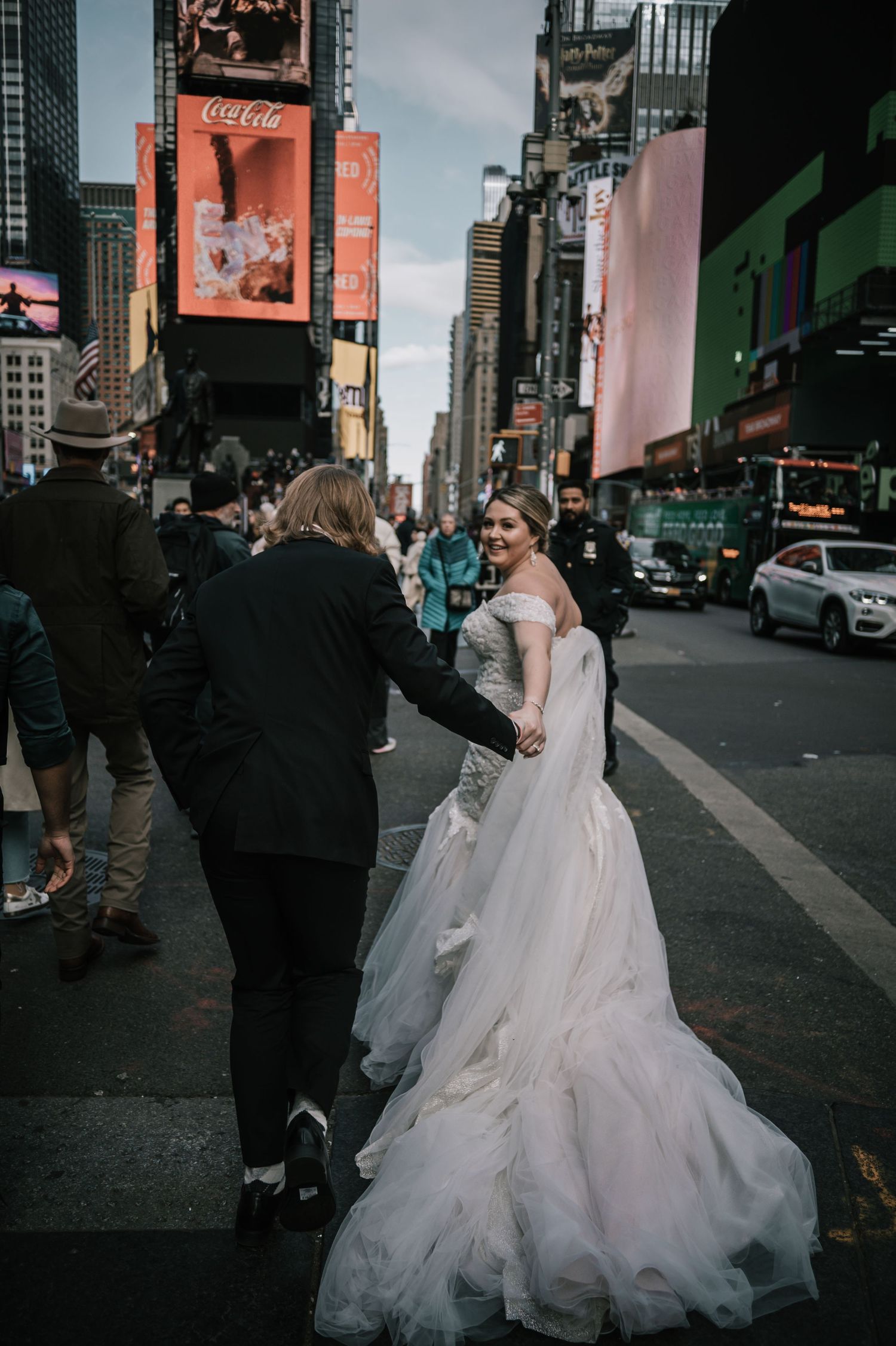 Newlyweds holding hands in Times Square NYC, bride in elegant white gown smiling at groom in black tuxedo.
