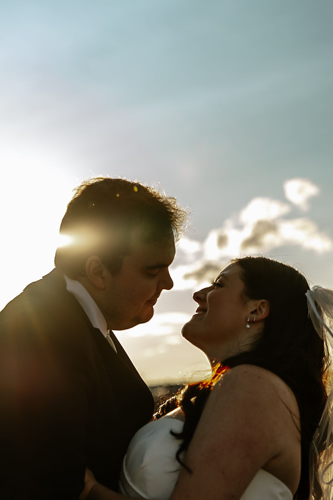 Groom kissing bride on cheek as she laughs in golden sunset light.
