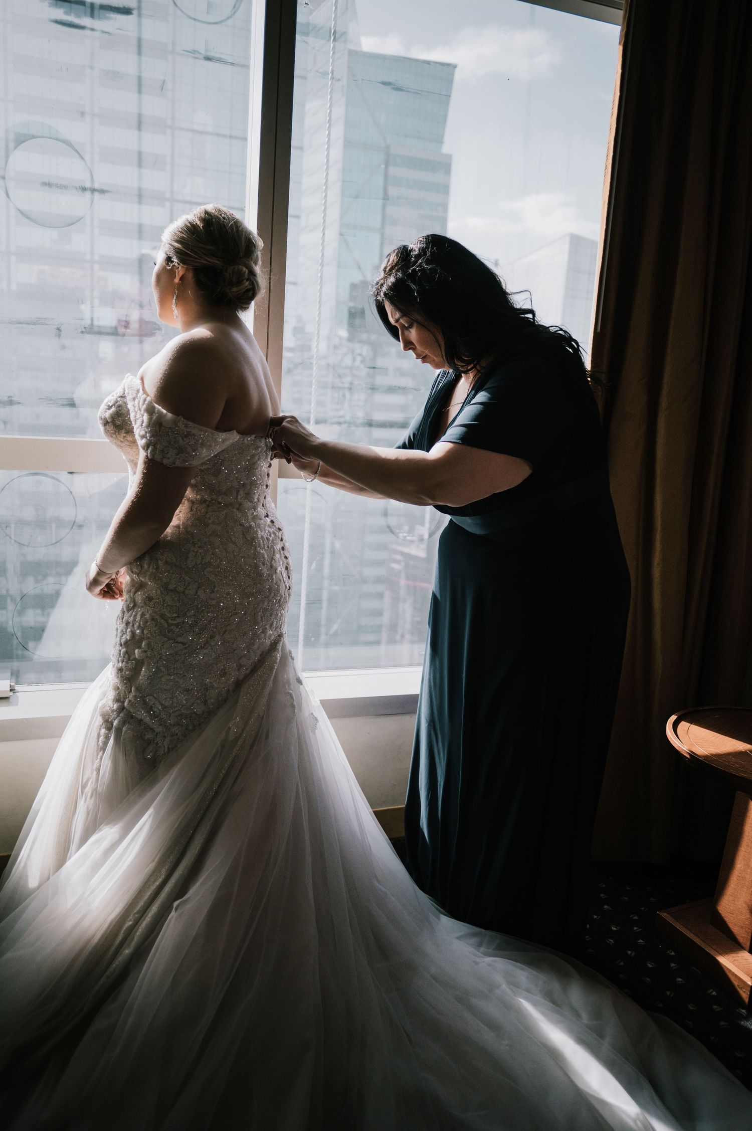Woman in teal dress fastens buttons on bride's lace mermaid wedding gown by city-view window.