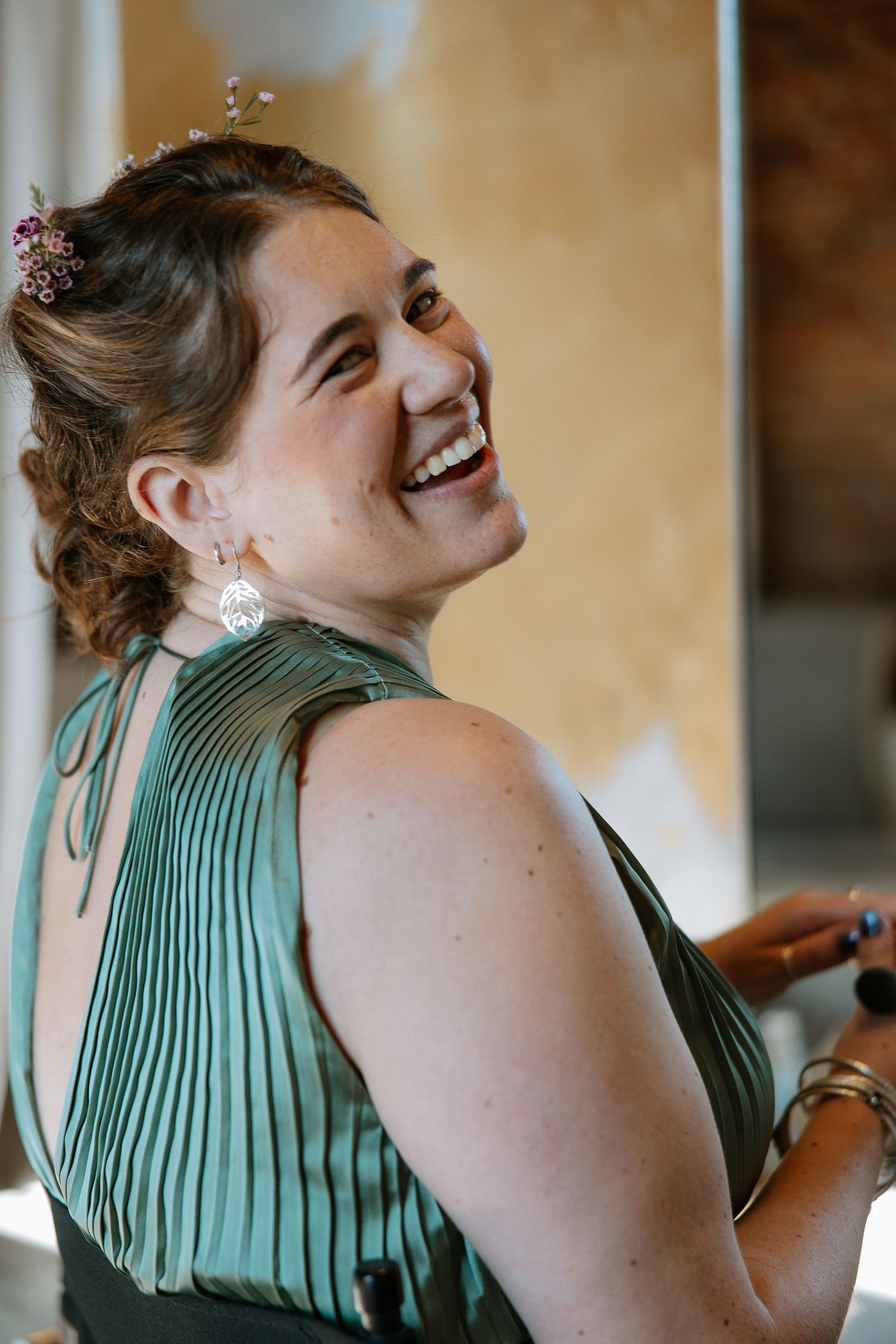 Smiling bridesmaid in green pleated dress with floral hair accessory and dangling earrings at a wedding celebration.