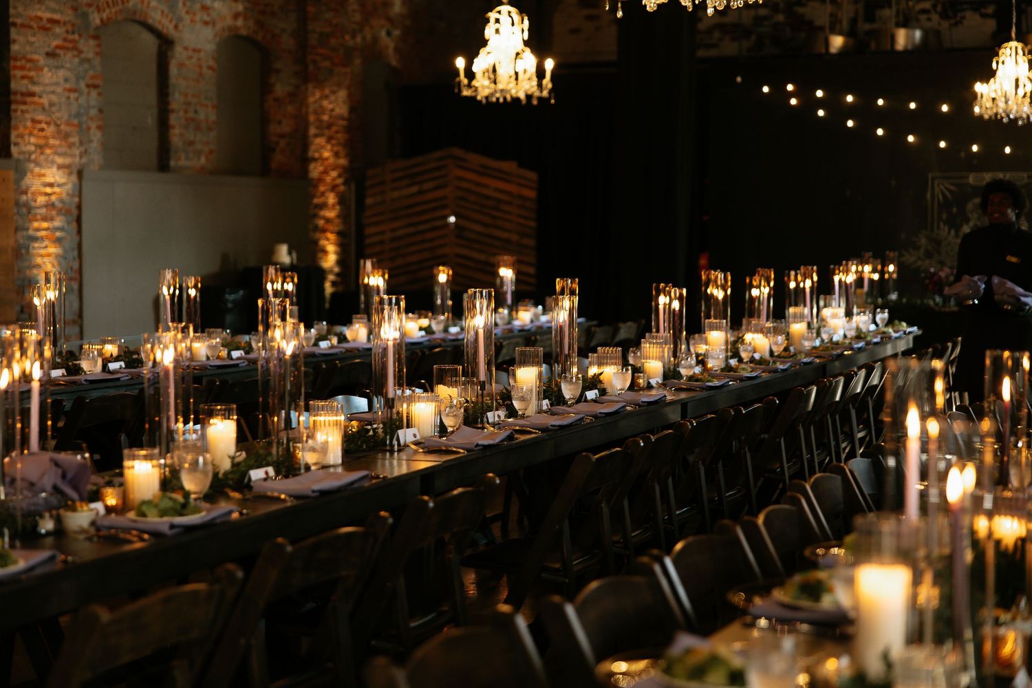 Elegant wedding reception with long banquet tables adorned with candles and glassware in a rustic brick venue.