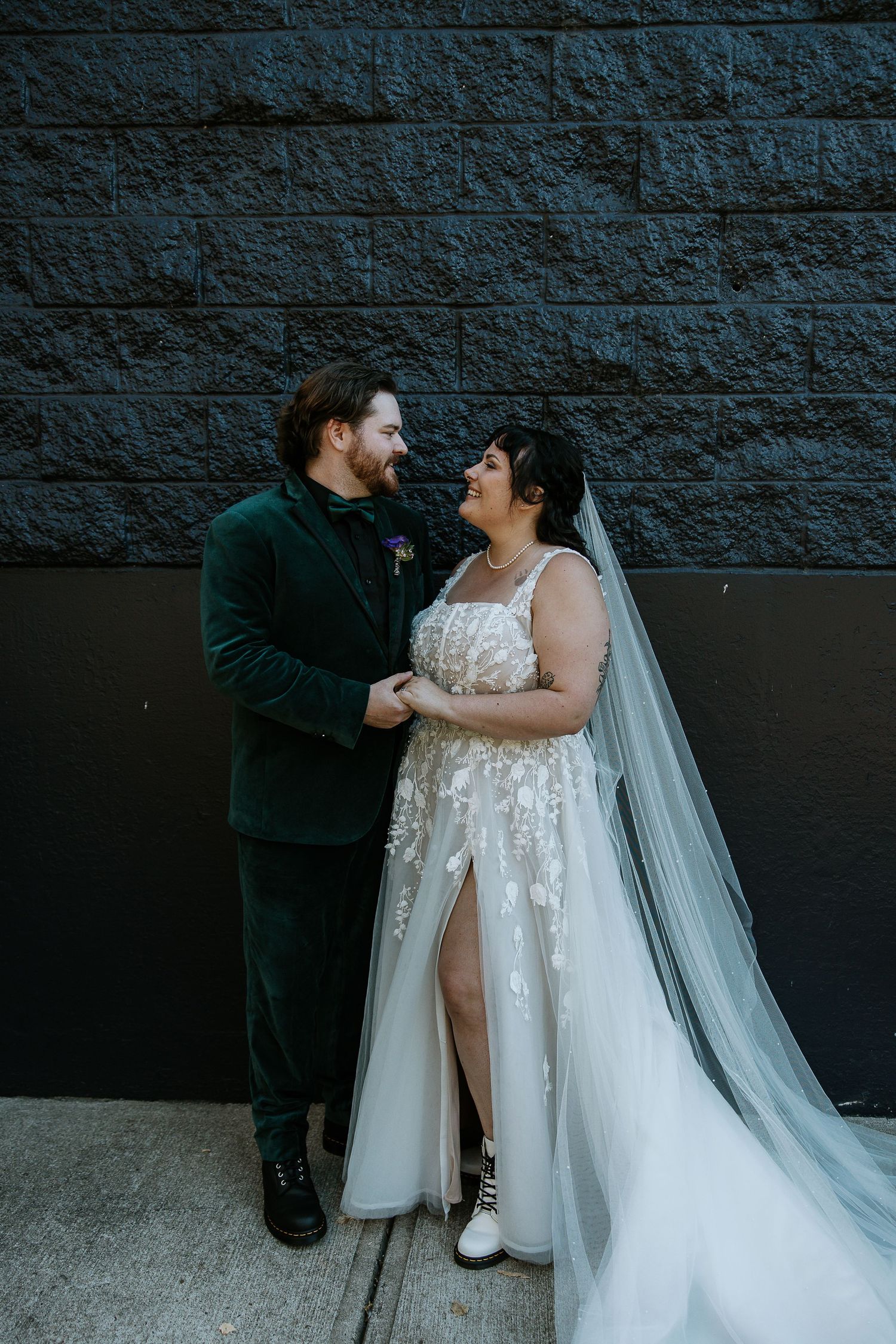 Bride and groom gaze lovingly at each other holding hands against dark textured stone wall outdoors.
