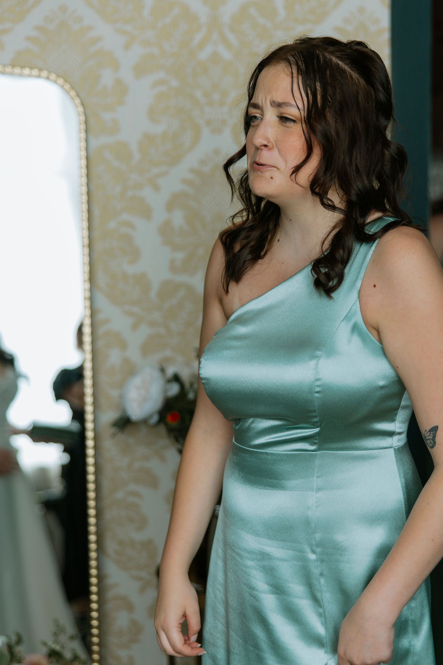 Bridesmaid in a satin mint green one-shoulder dress standing near a gold-framed mirror.