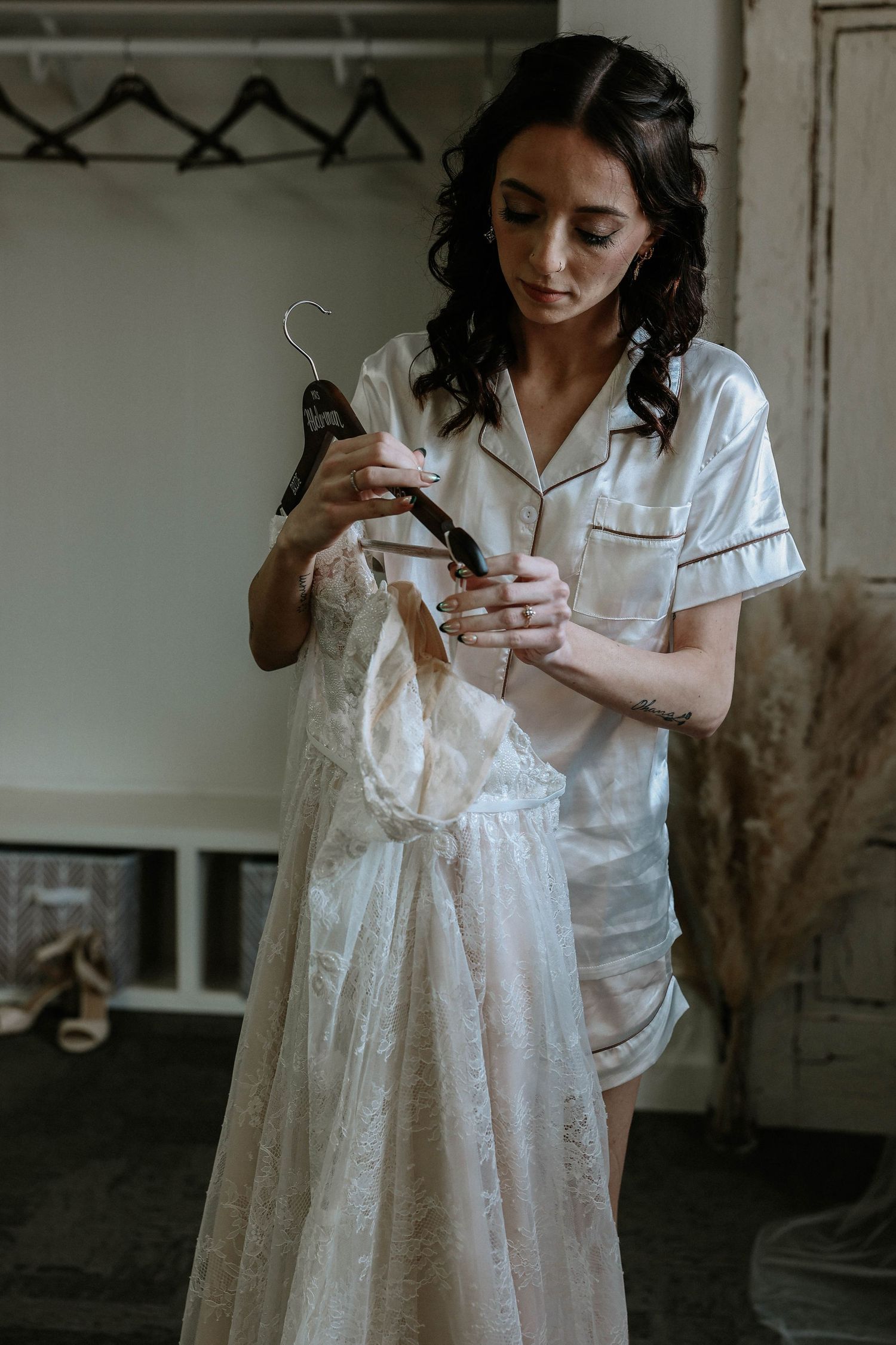 Bride in white satin pajamas holding a lace wedding dress on a hanger in a softly lit dressing room.