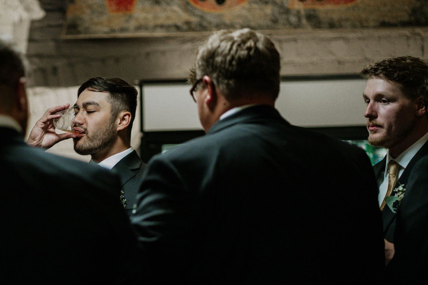 Groom drinking a shot with groomsmen in dark suits before wedding ceremony indoors.