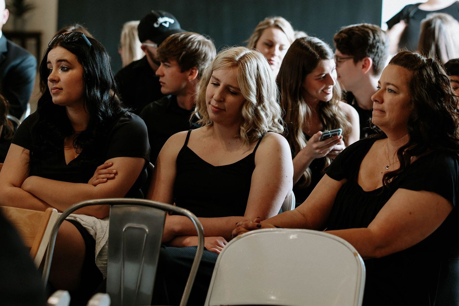 Group of young women in black outfits seated in white chairs at an indoor event, engaged in conversation.