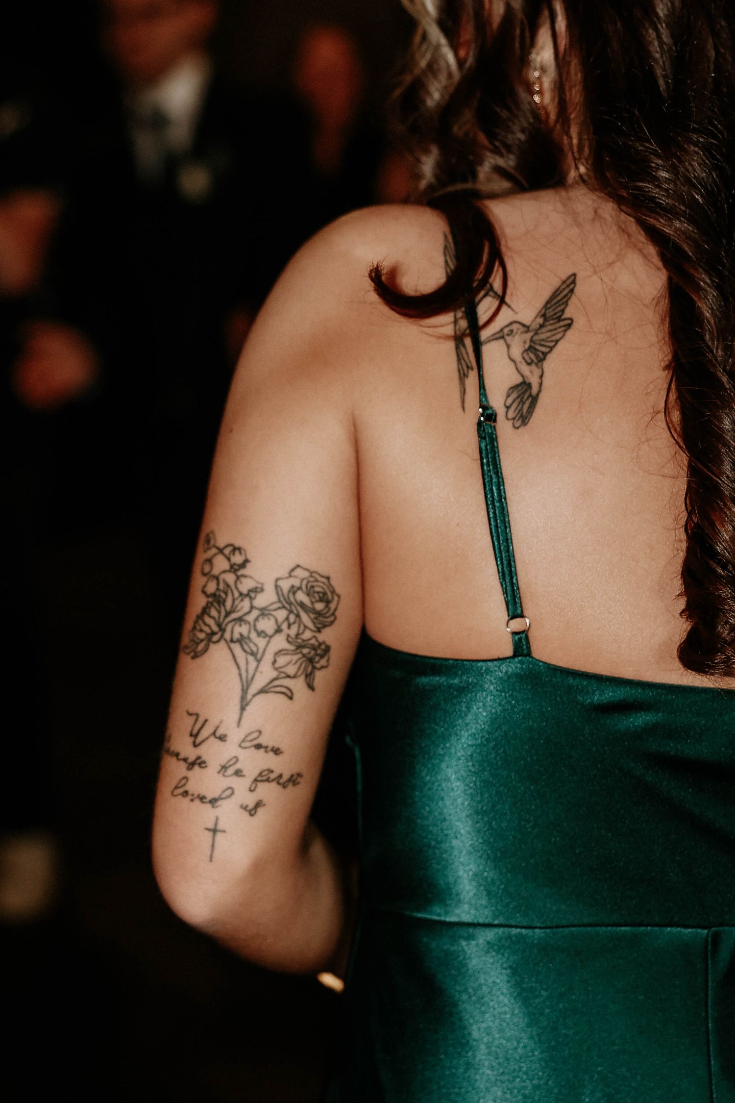 Woman in green satin dress with hummingbird back tattoo and floral arm tattoo with script at a formal event.