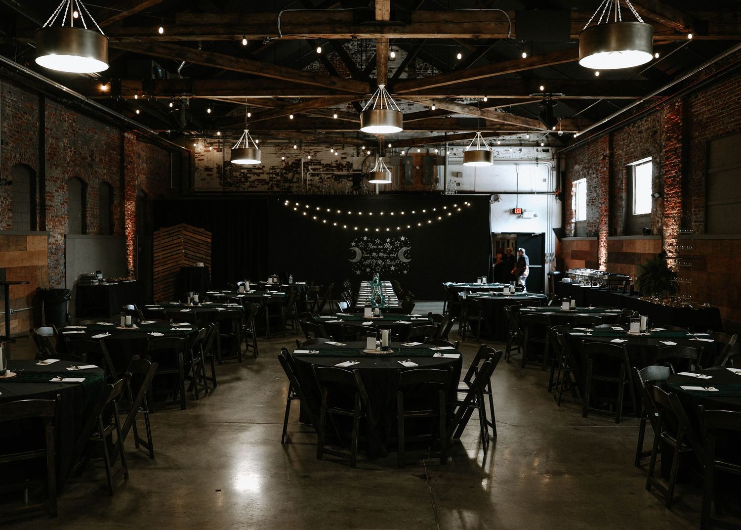 Dark industrial event venue with black tables, string lights, and exposed brick walls set for a formal dinner.