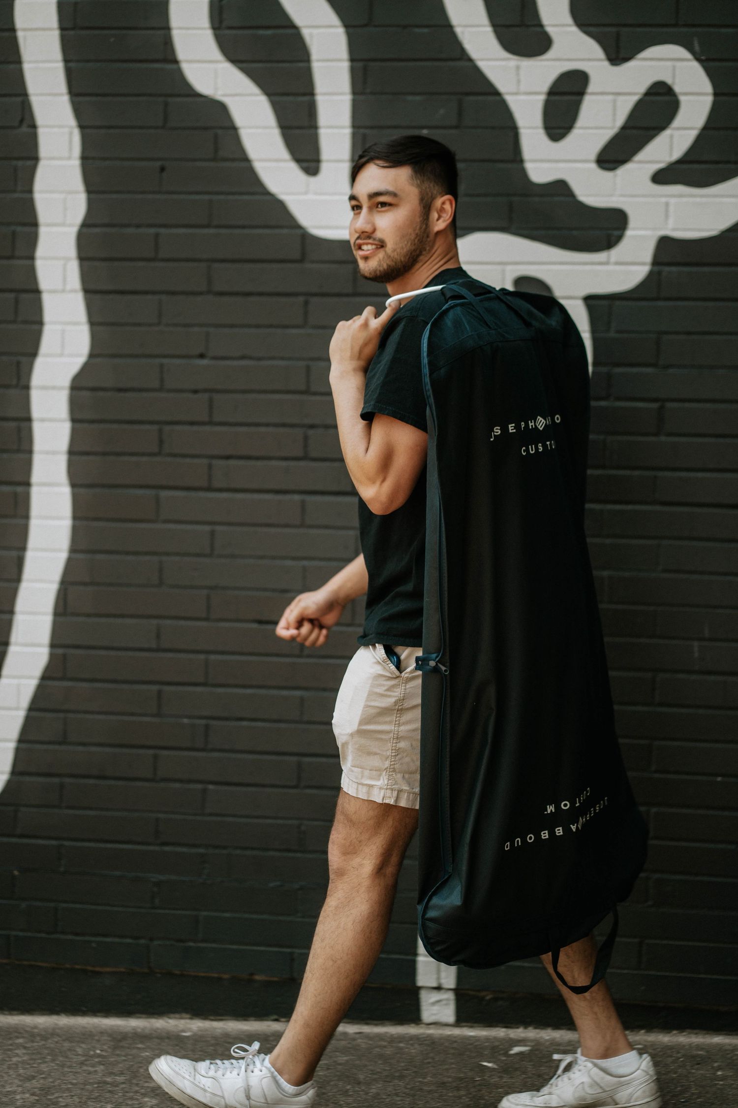 Young man carrying a black garment bag over shoulder walking past a graffiti brick wall outdoors.