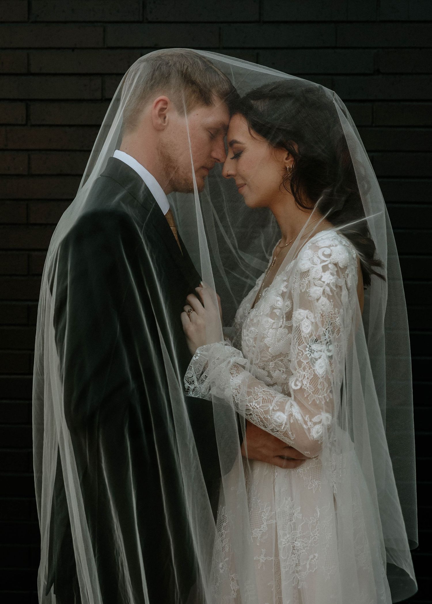 Bride and groom share an intimate moment under a sheer wedding veil against a dark brick wall.