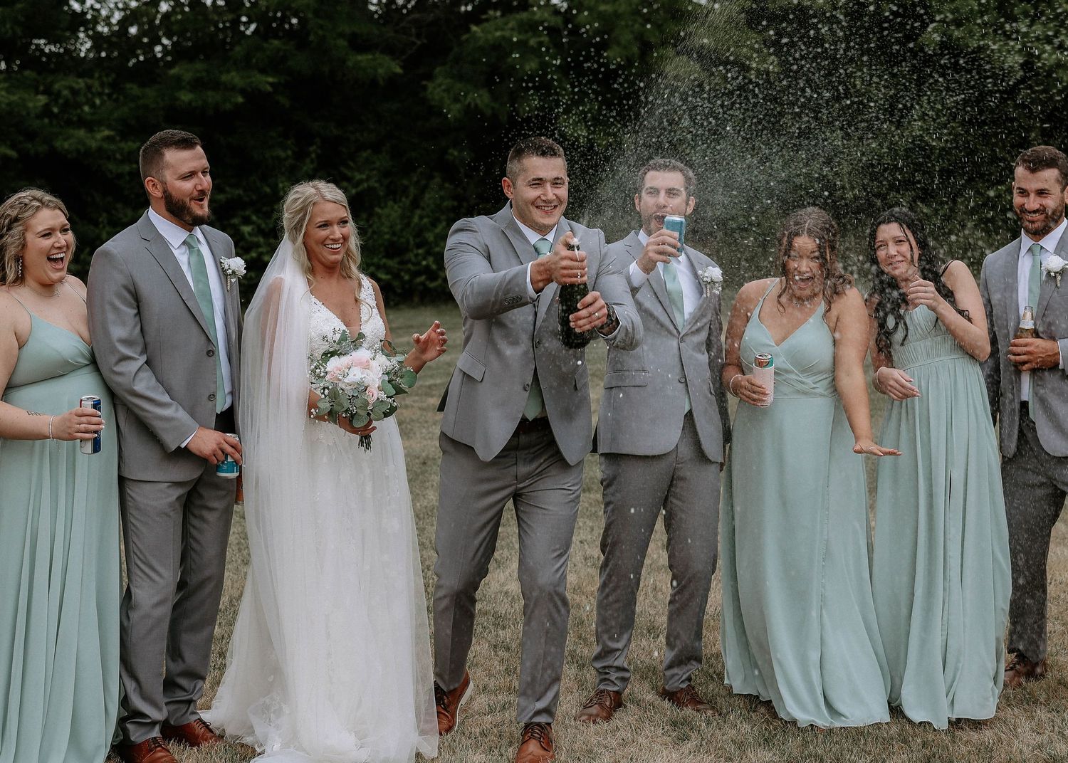 Wedding party celebrates with champagne spray outdoors, bridesmaids in sage green gowns and groomsmen in gray suits.