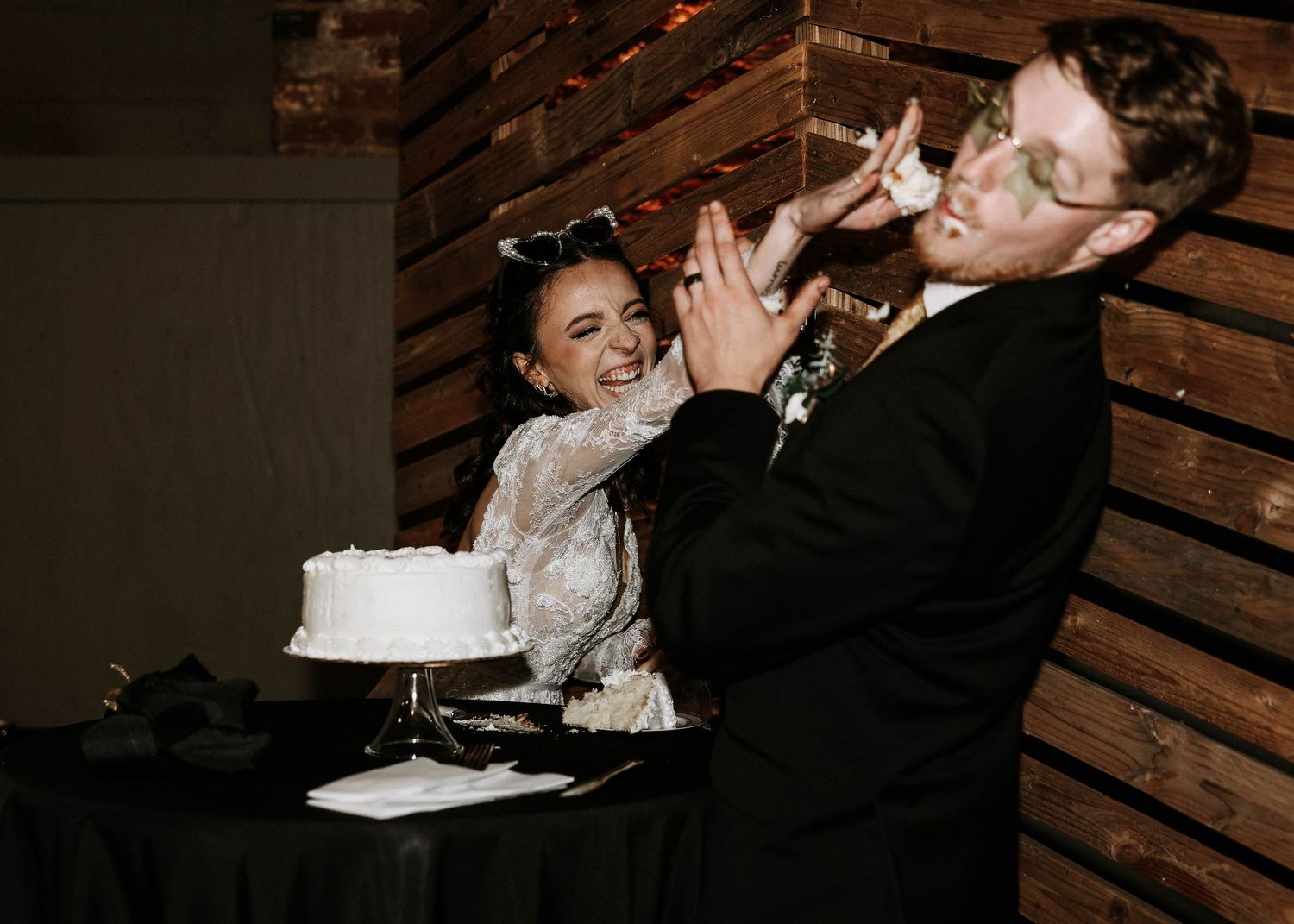Bride in lace gown playfully smashes wedding cake into grooms face during reception near white cake on table.