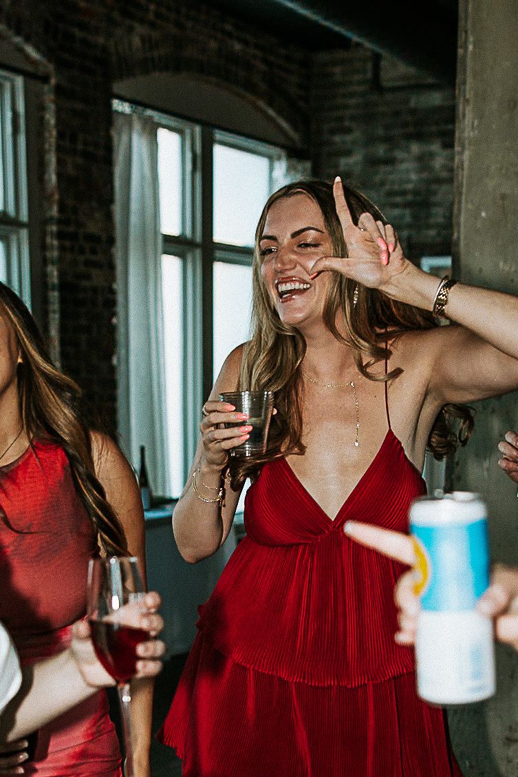 Woman in red dress smiles and dances joyfully while holding a drink at a wedding reception.