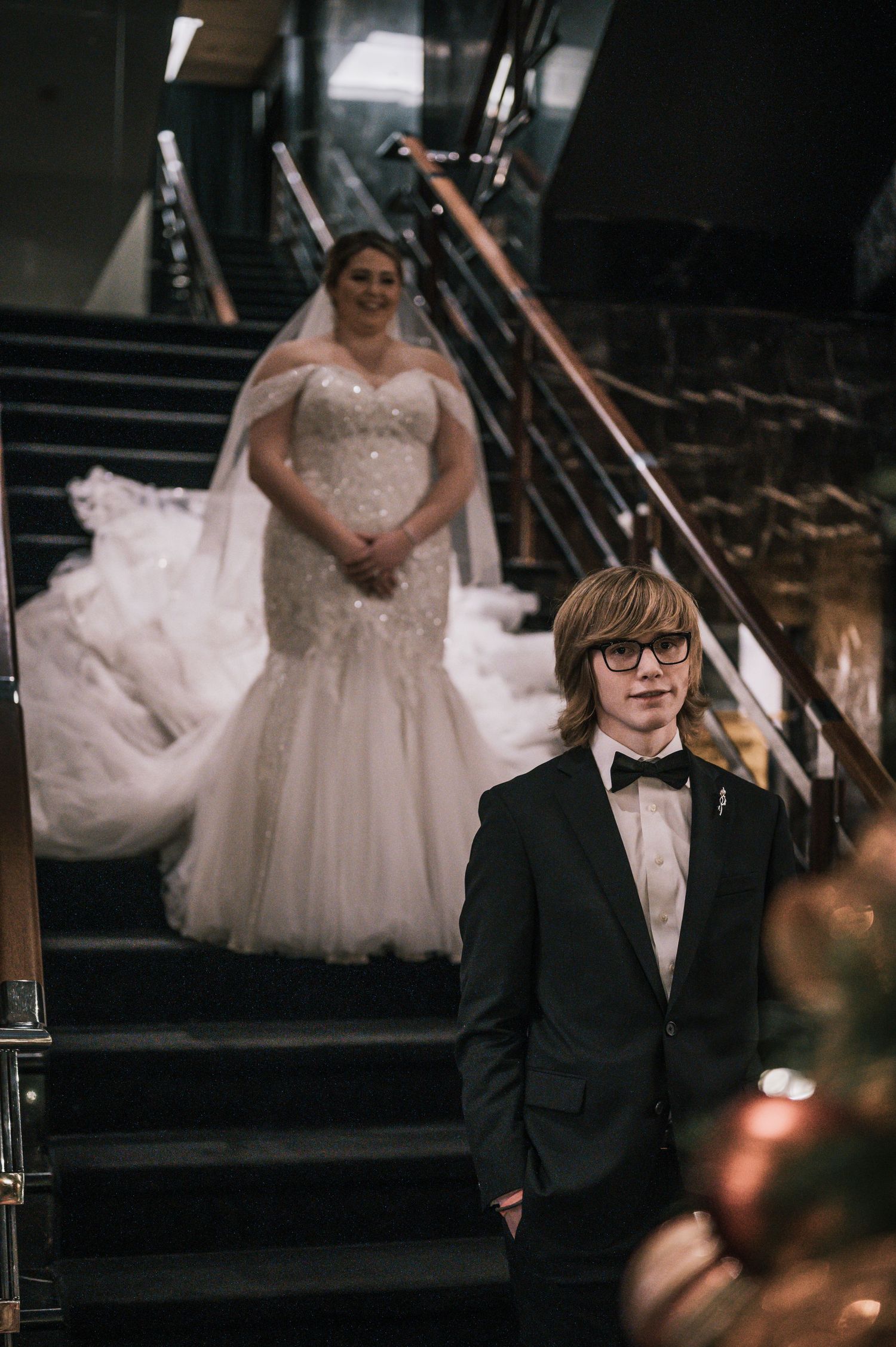 Young man in tuxedo stands on staircase as bride in beaded mermaid gown descends behind him.