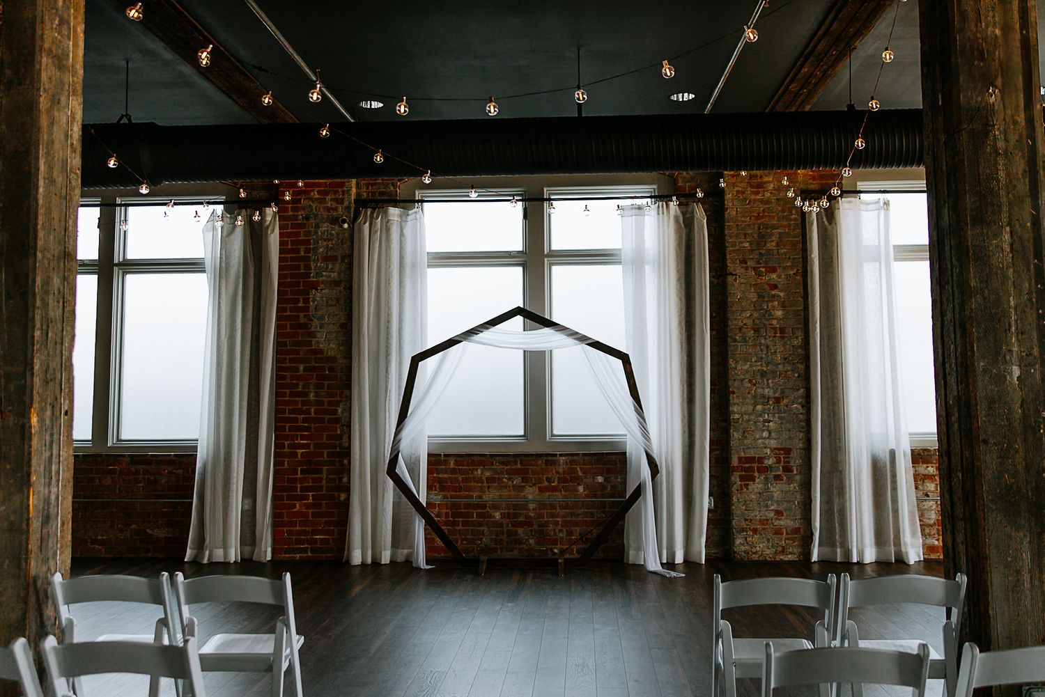 Elegant wedding ceremony venue with hexagonal arch, white drapes, brick walls, string lights, and white chairs.