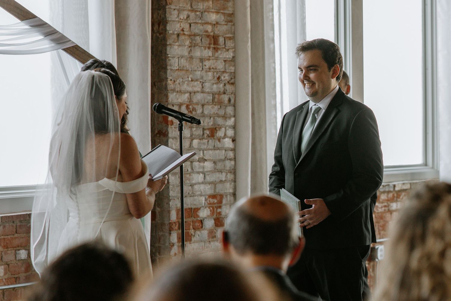 Smiling groom in black suit listens as bride reads wedding vows at altar.