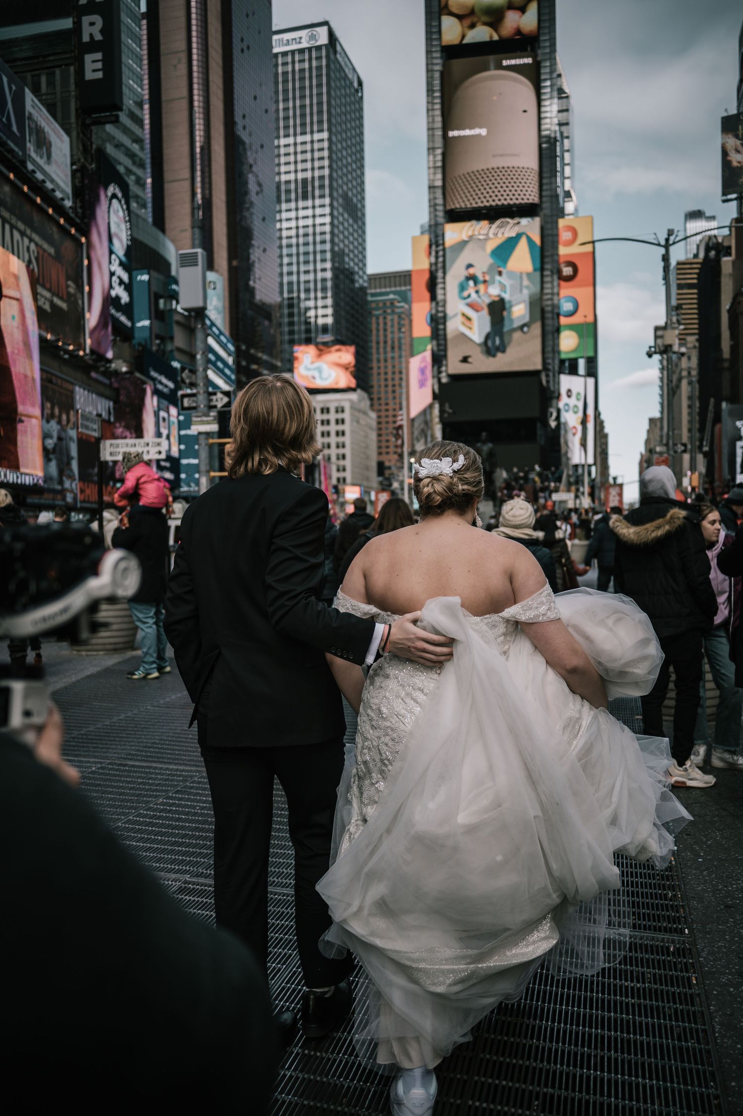 Bride and groom walking through Times Square NYC, bride in white gown holding dress train amid city crowds.