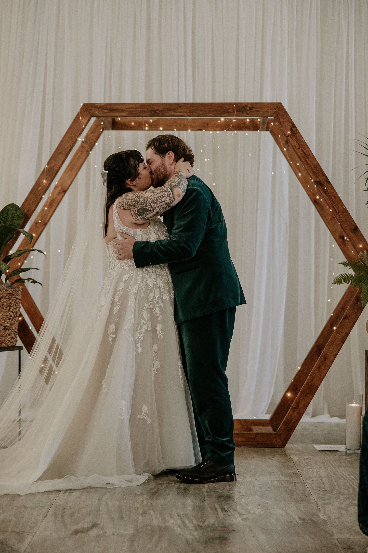 Bride and groom share first kiss under hexagonal wooden arch with fairy lights backdrop.