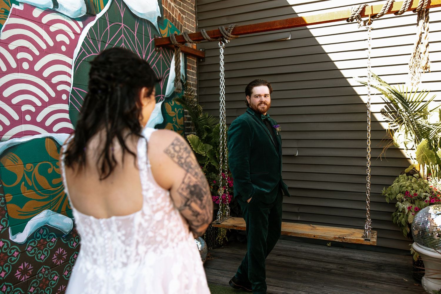 Groom in green velvet suit smiling at tattooed bride in lace wedding dress during outdoor first look.