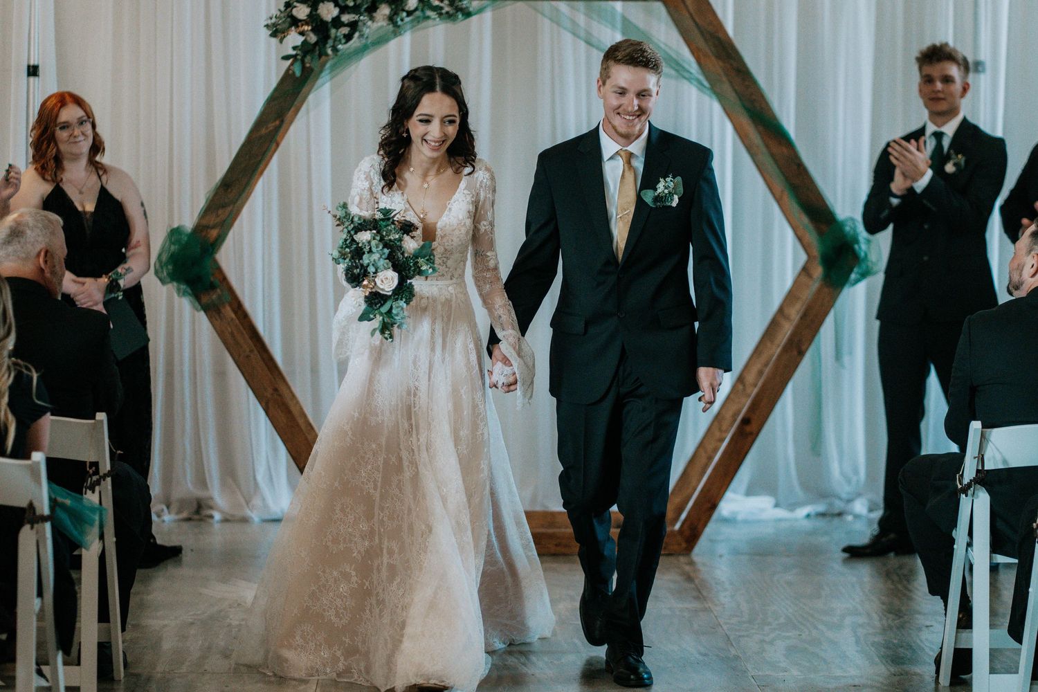 Bride and groom walk down the aisle after wedding ceremony beneath a geometric wooden hexagon arch with green floral decor.