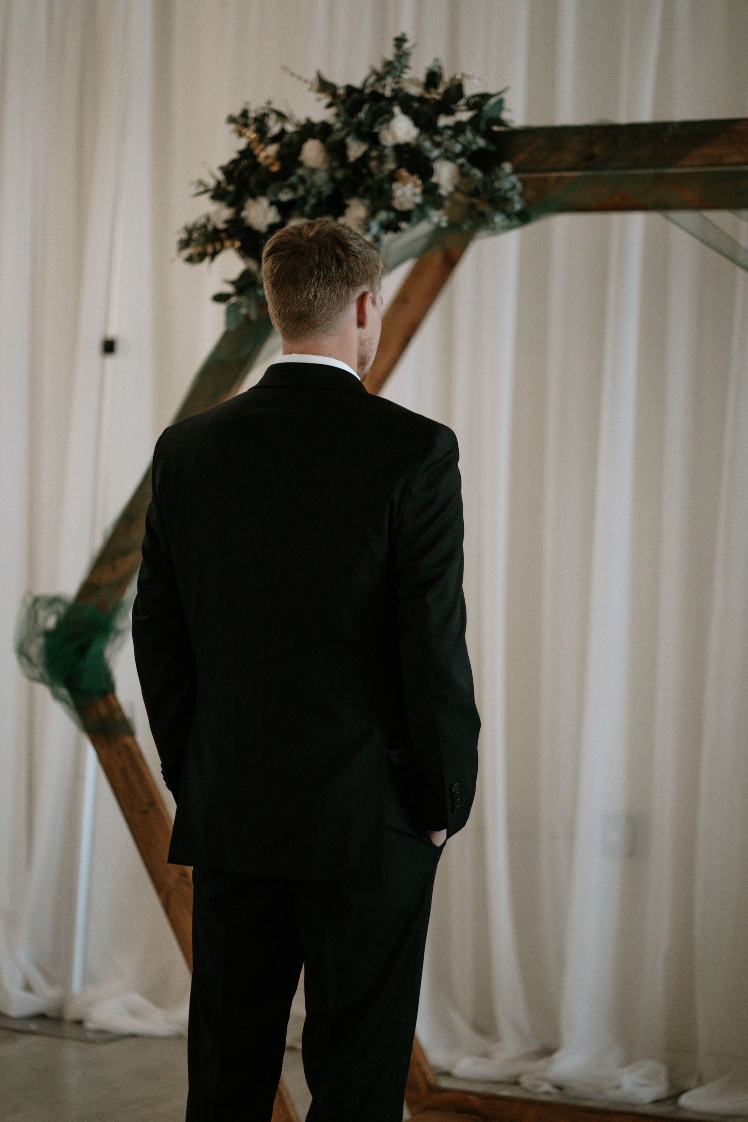 Groom in black suit standing at wooden geometric wedding arch decorated with white roses and greenery.