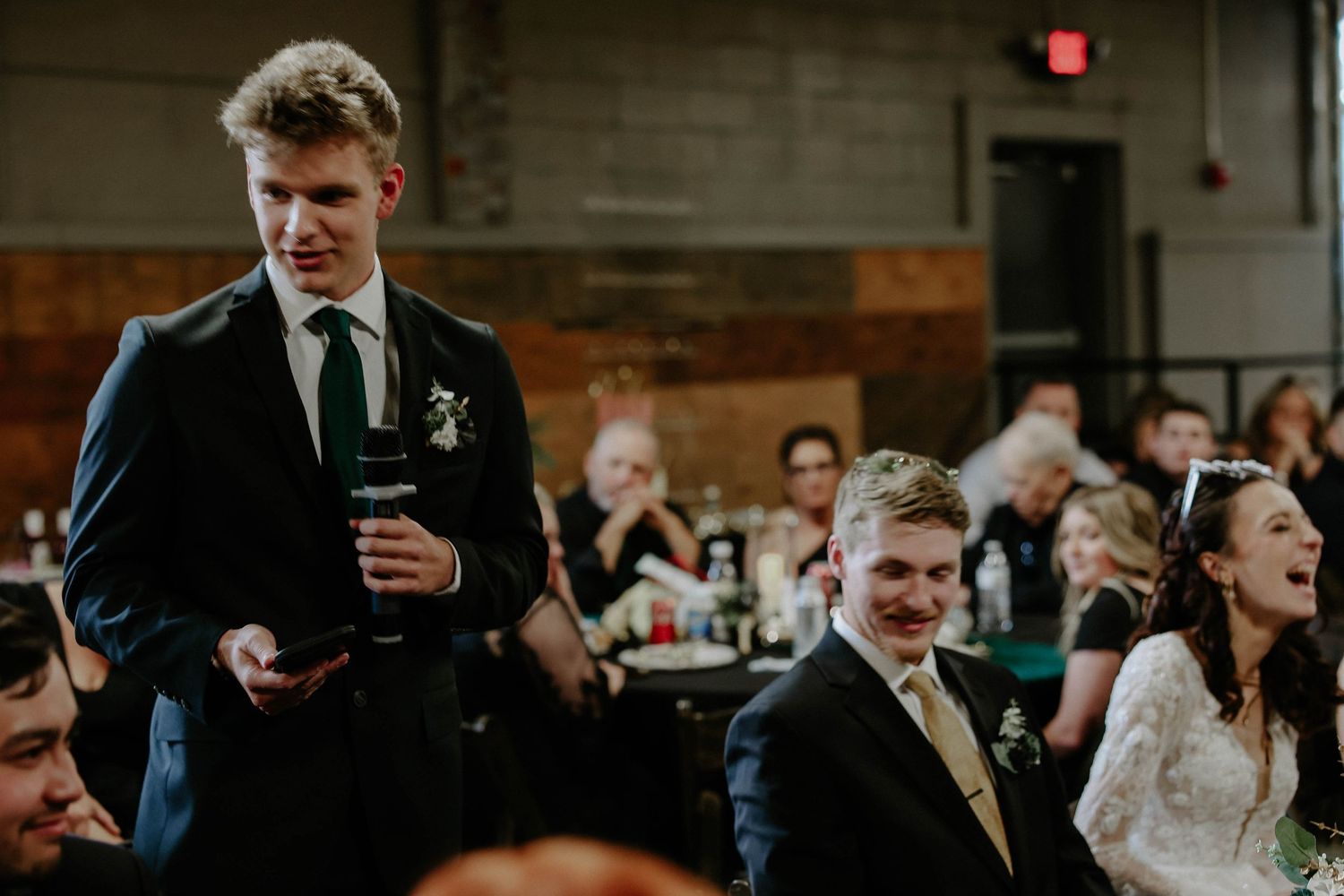 Young man in navy suit giving a wedding toast speech while guests laugh and celebrate at a reception.
