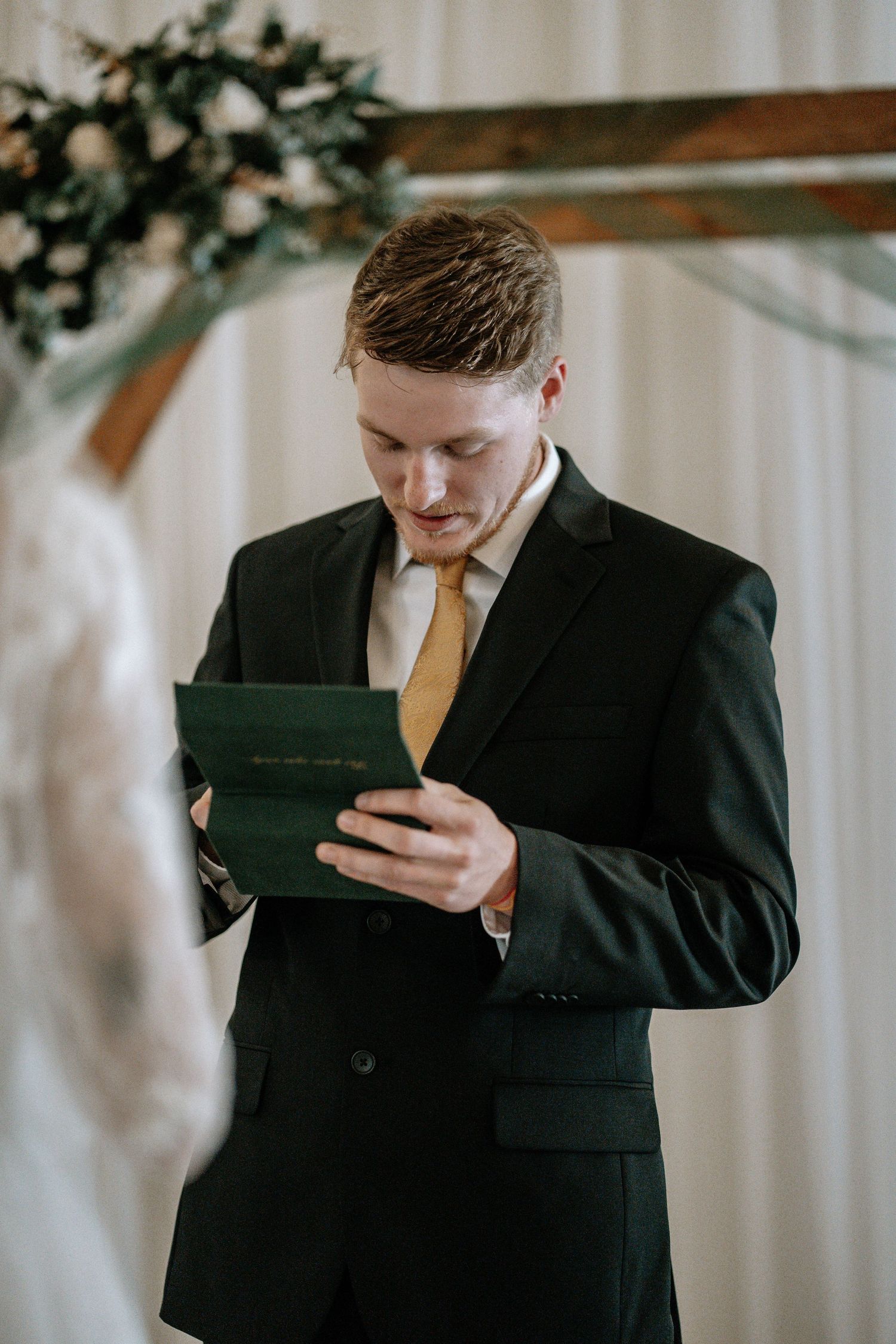 Groom in black suit and gold tie reads wedding vows from green booklet at altar decorated with floral arch.