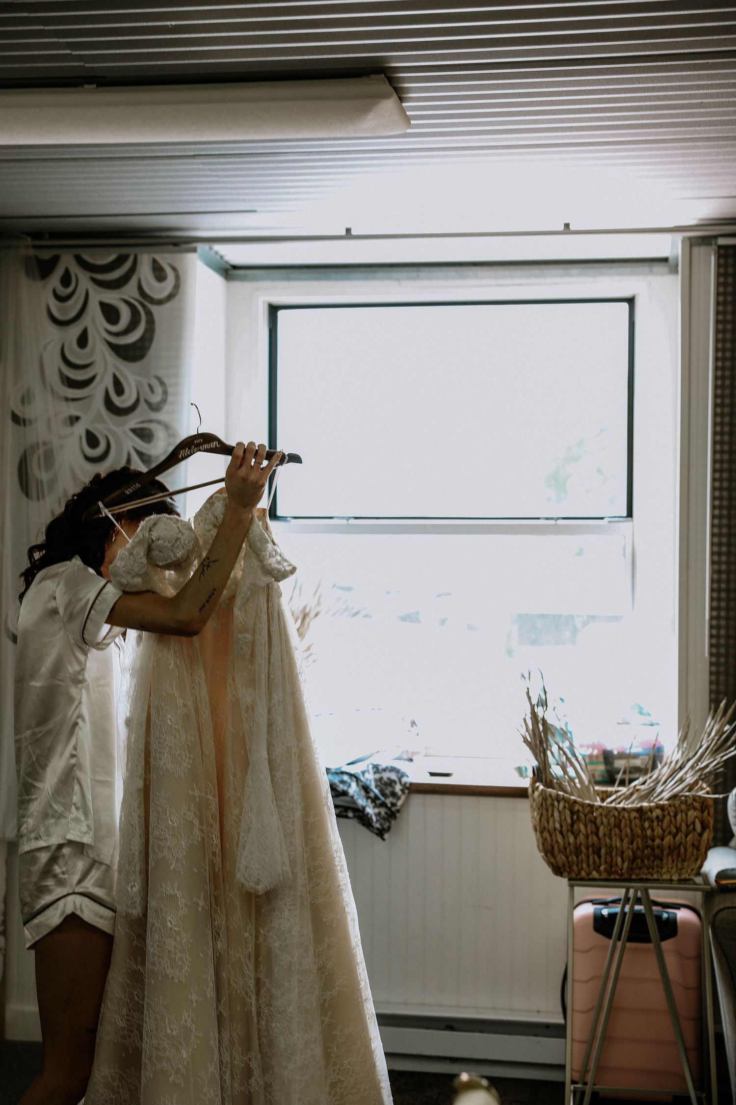 Elegant lace wedding dress hanging near bright window in a cozy bridal preparation room.