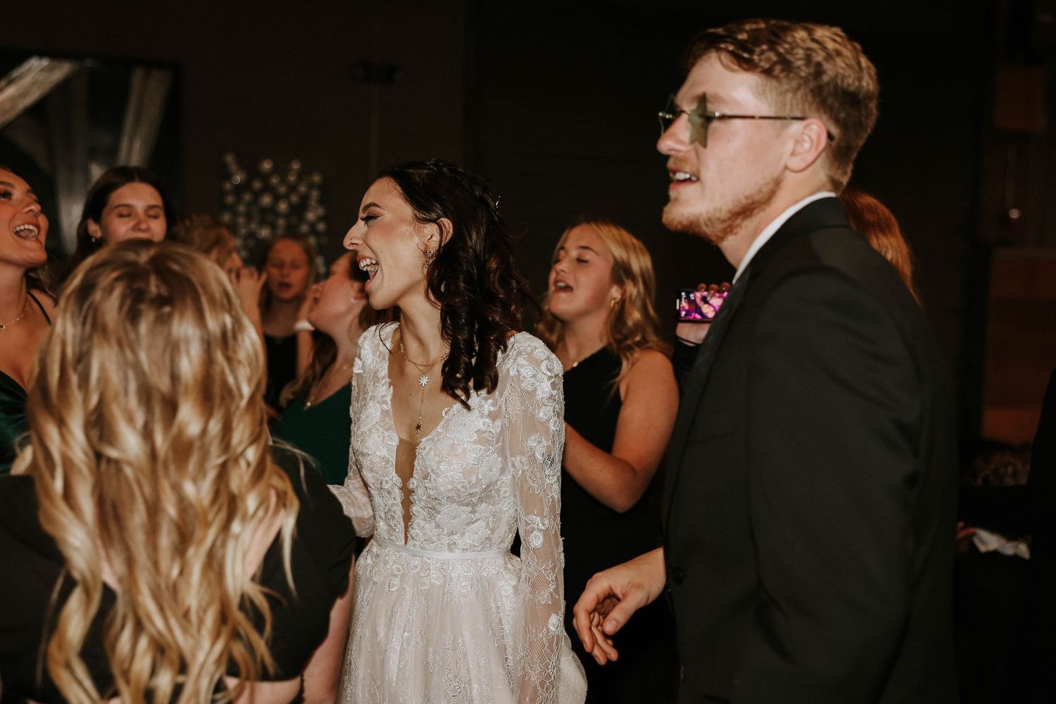 Bride in lace gown and groom in black suit singing joyfully with guests at wedding reception dance floor.