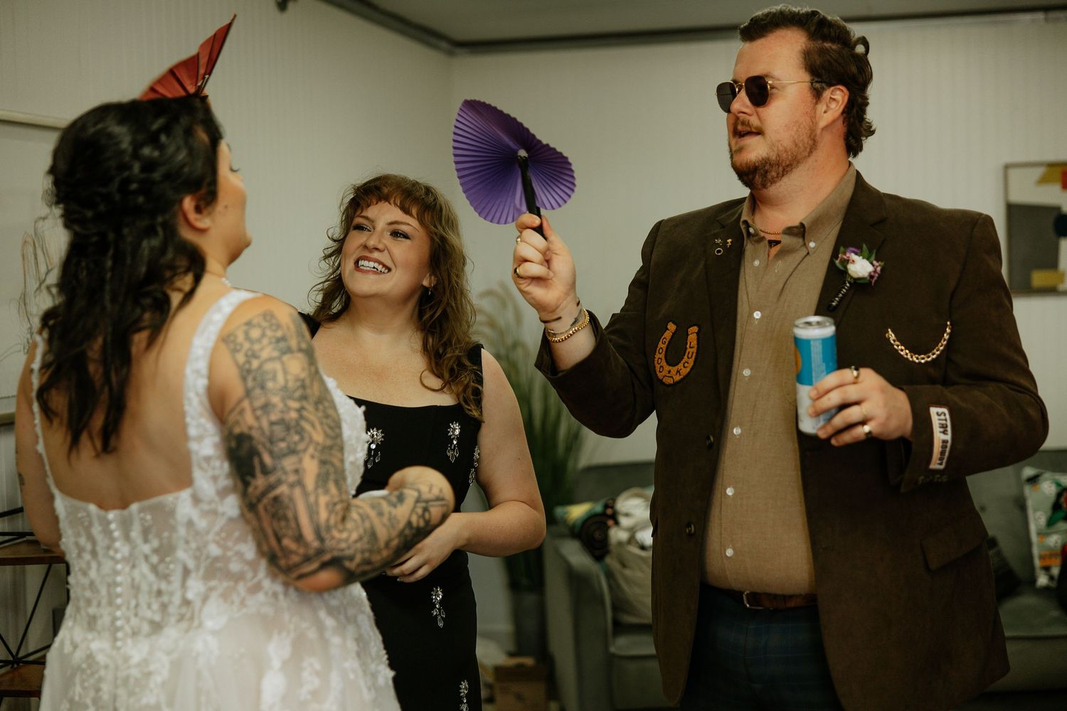 Bride in white lace dress laughs with tattooed woman and man in brown jacket holding purple fan at wedding reception.