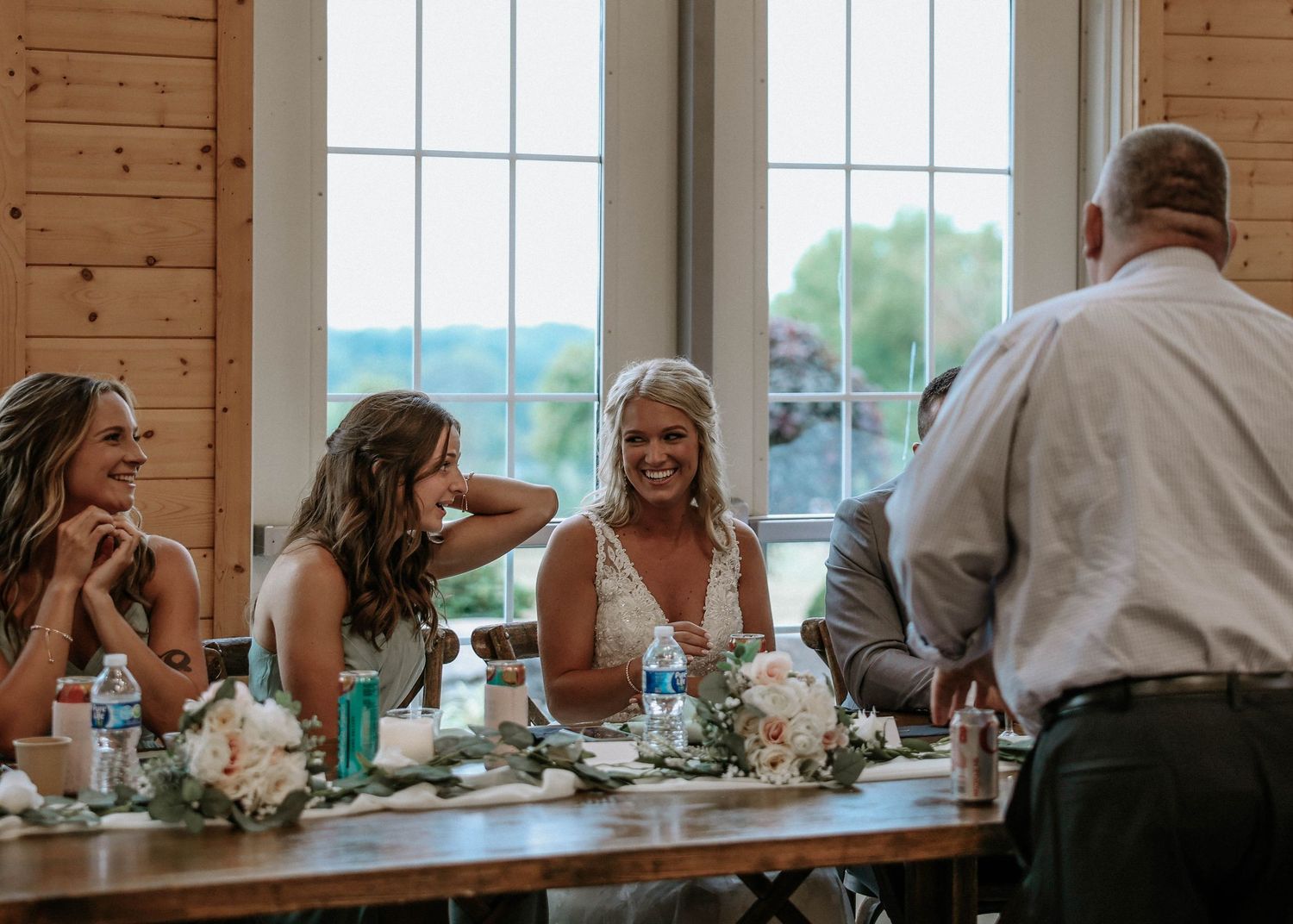 Bride laughing joyfully with bridesmaids at rustic wooden wedding reception table adorned with floral centerpieces.
