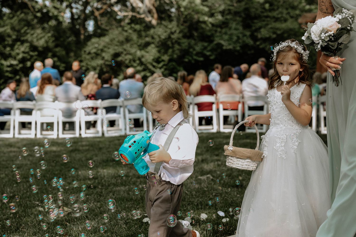 Ring bearer and flower girl walk down the outdoor wedding aisle, with bubbles floating around them during the ceremony.