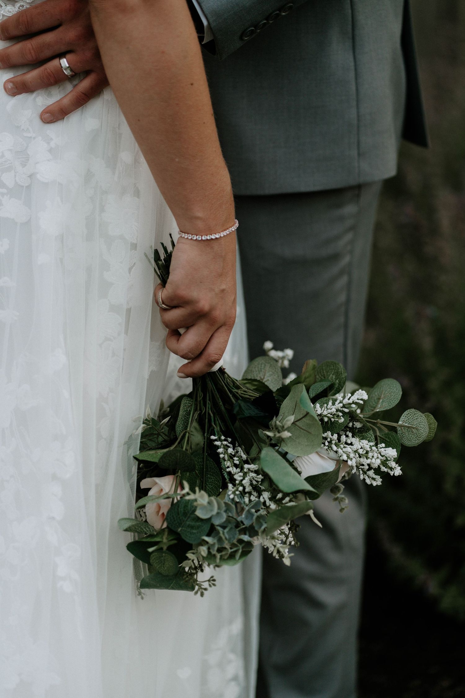 Bride and groom holding hands with a green eucalyptus wedding bouquet, bride in lace dress and groom in gray suit.