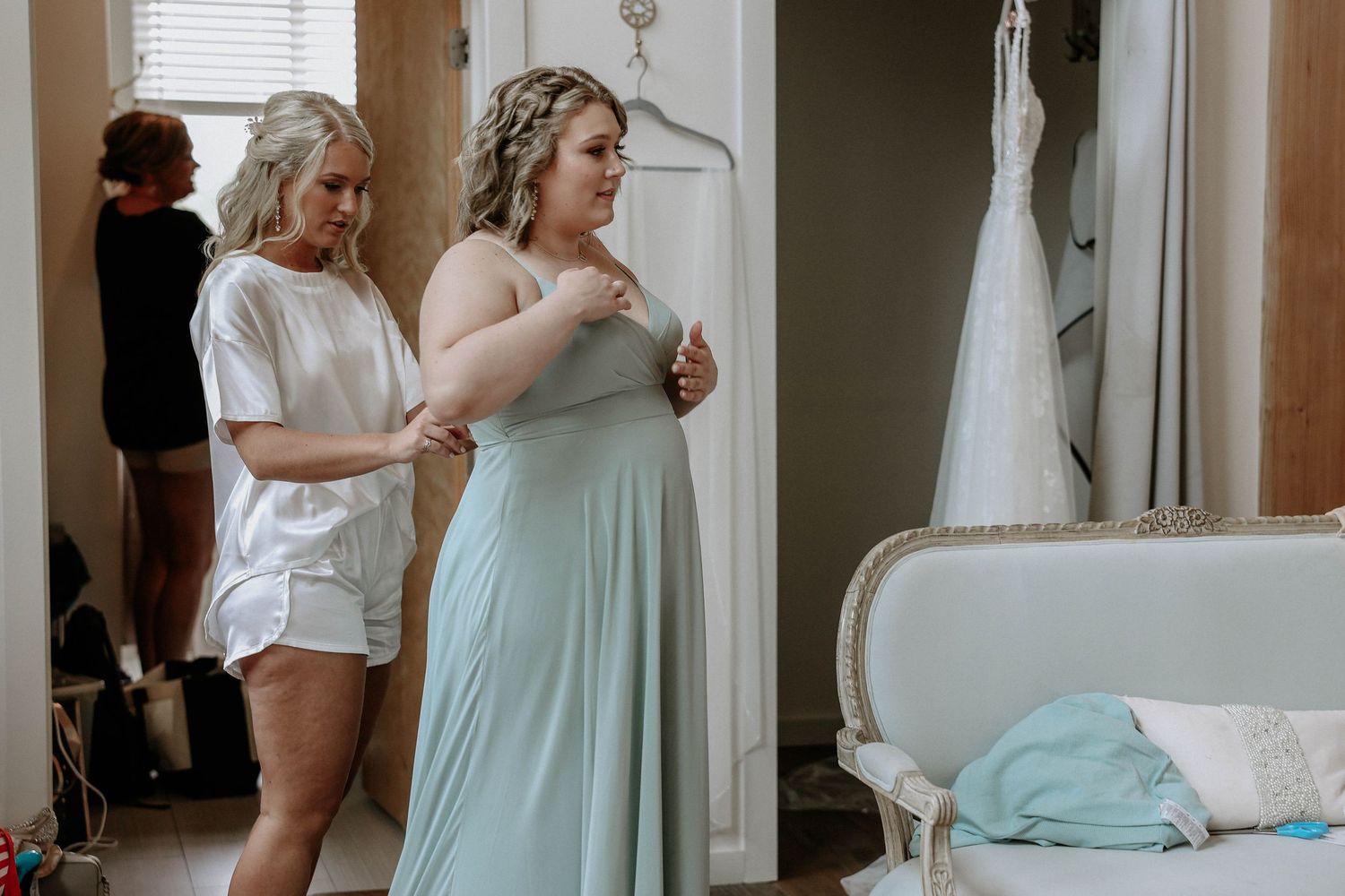 Bridesmaid in mint green gown being helped with dress while wedding gown hangs in background.