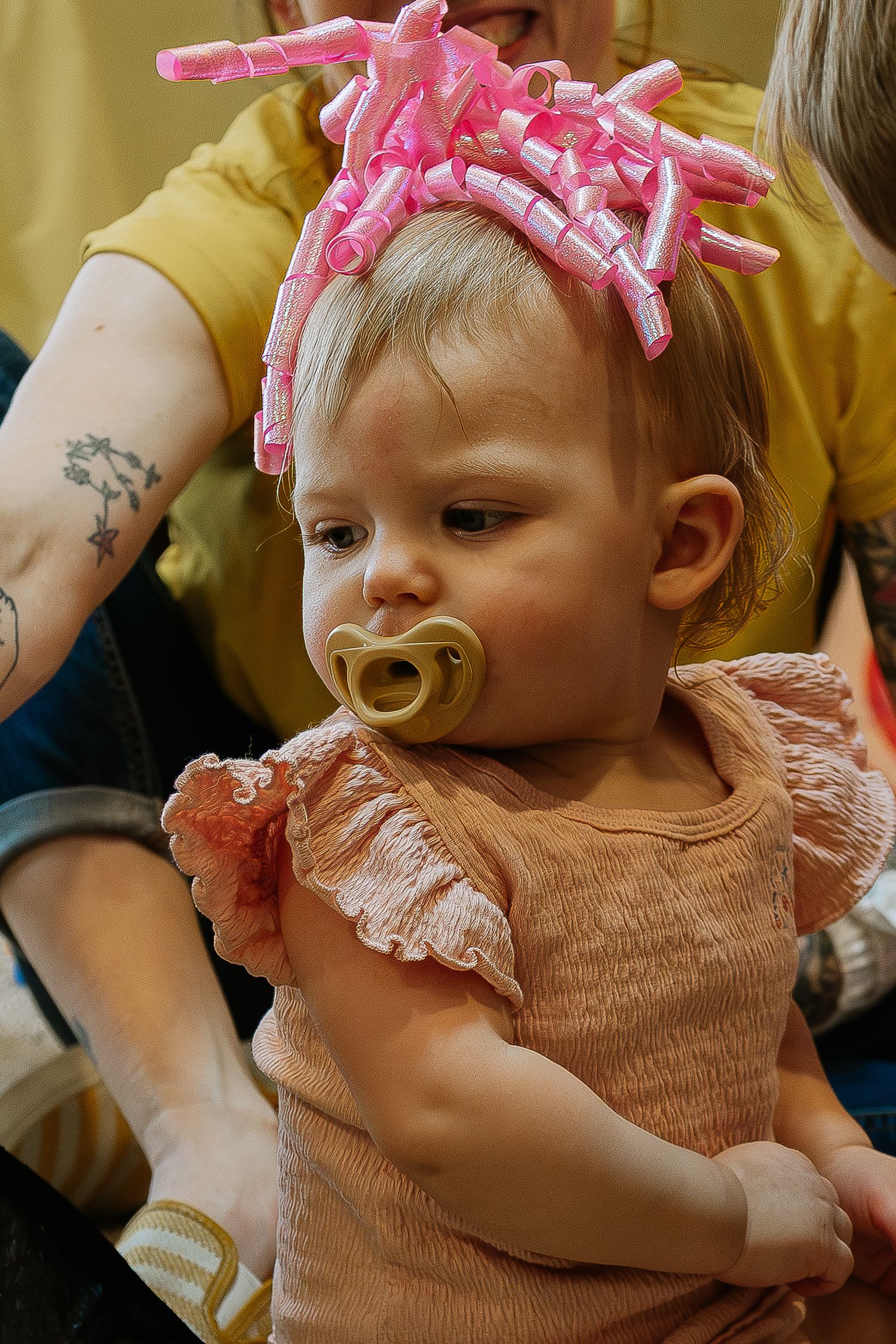 Adorable baby wearing pink curler headband and pacifier in peach ruffled dress being held by adult in Dayton, Ohio