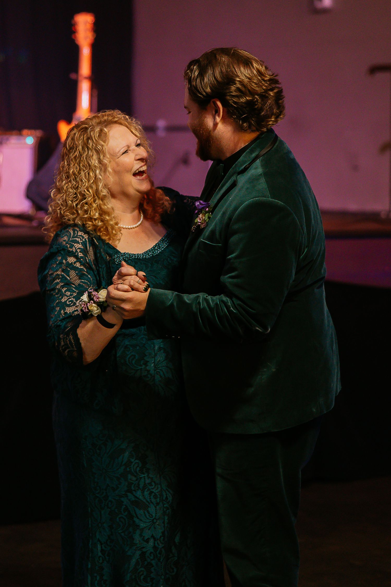 A groom in a green velvet suit shares a mother-son dance at a wedding reception.