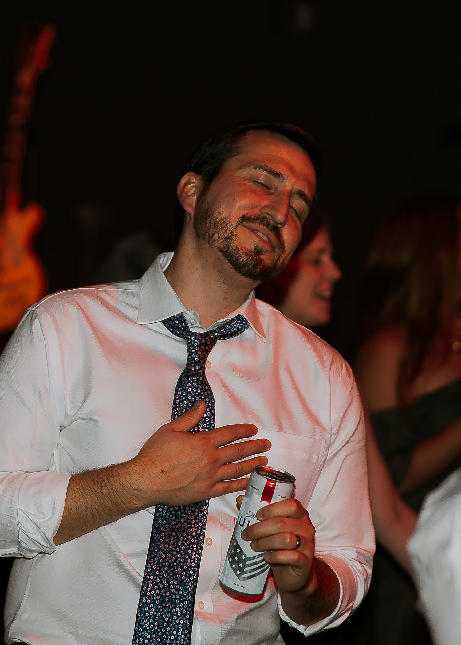 Man in white shirt and tie dances with eyes closed holding drink at party.