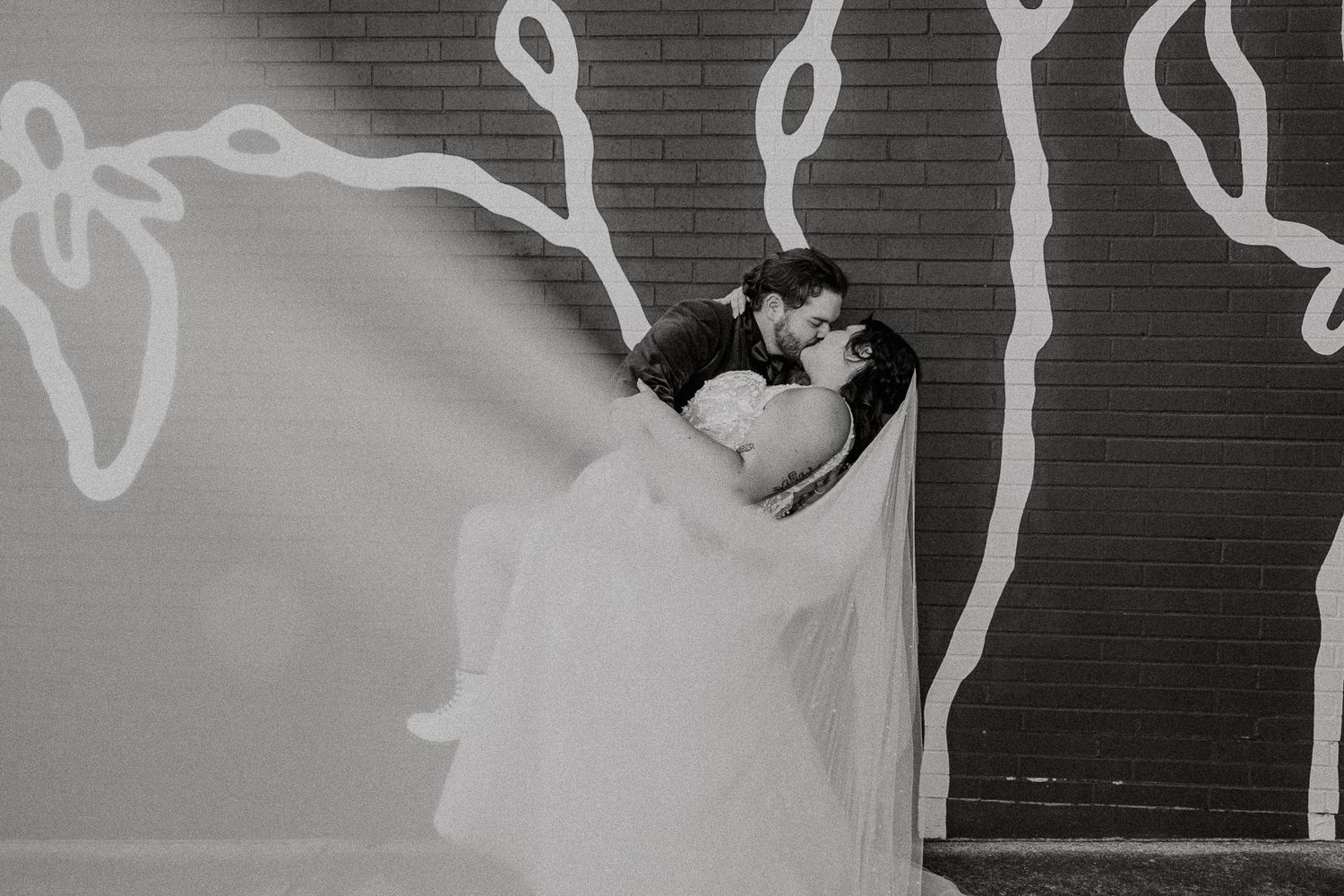 Black and white photo of bride and groom kissing against mural wall, veil flowing romantically.