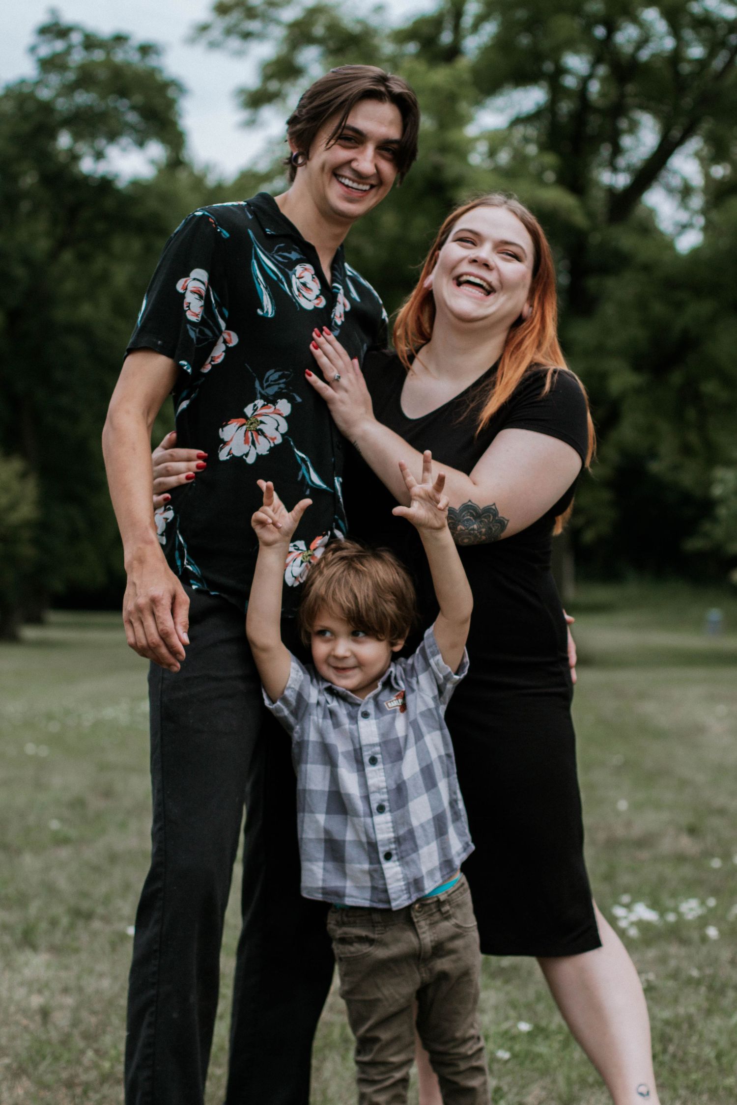 A joyful family of three posing outdoors in a park, with parents smiling and a young boy raising his arms at Eden Park in Cincinnati, Ohio