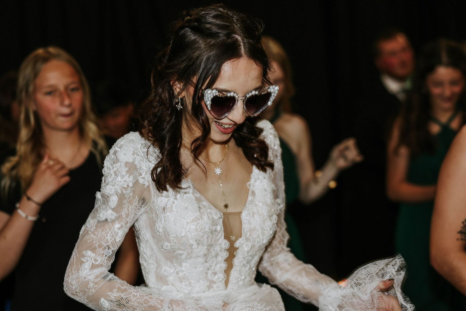 Bride in lace wedding dress and heart-shaped sunglasses dancing joyfully at wedding reception.