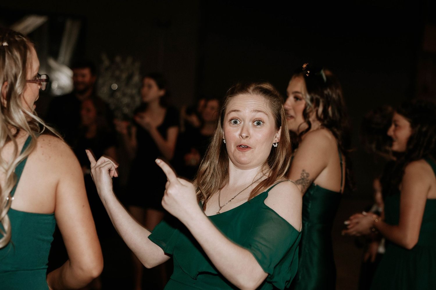 Woman in green off-shoulder dress dancing and pointing fingers at wedding reception party.