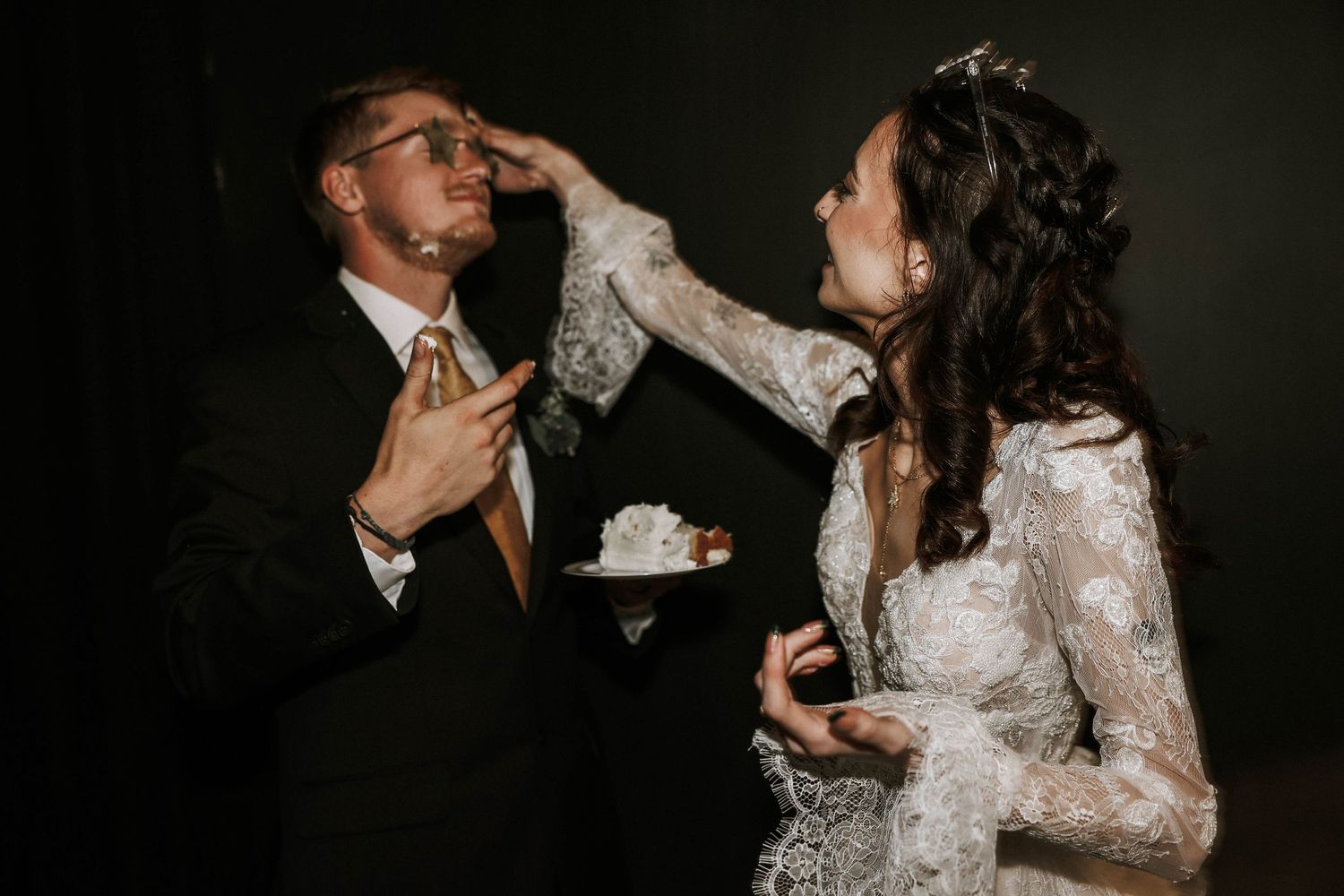 Bride in lace gown feeds wedding cake to groom in suit during fun cake cutting ceremony.