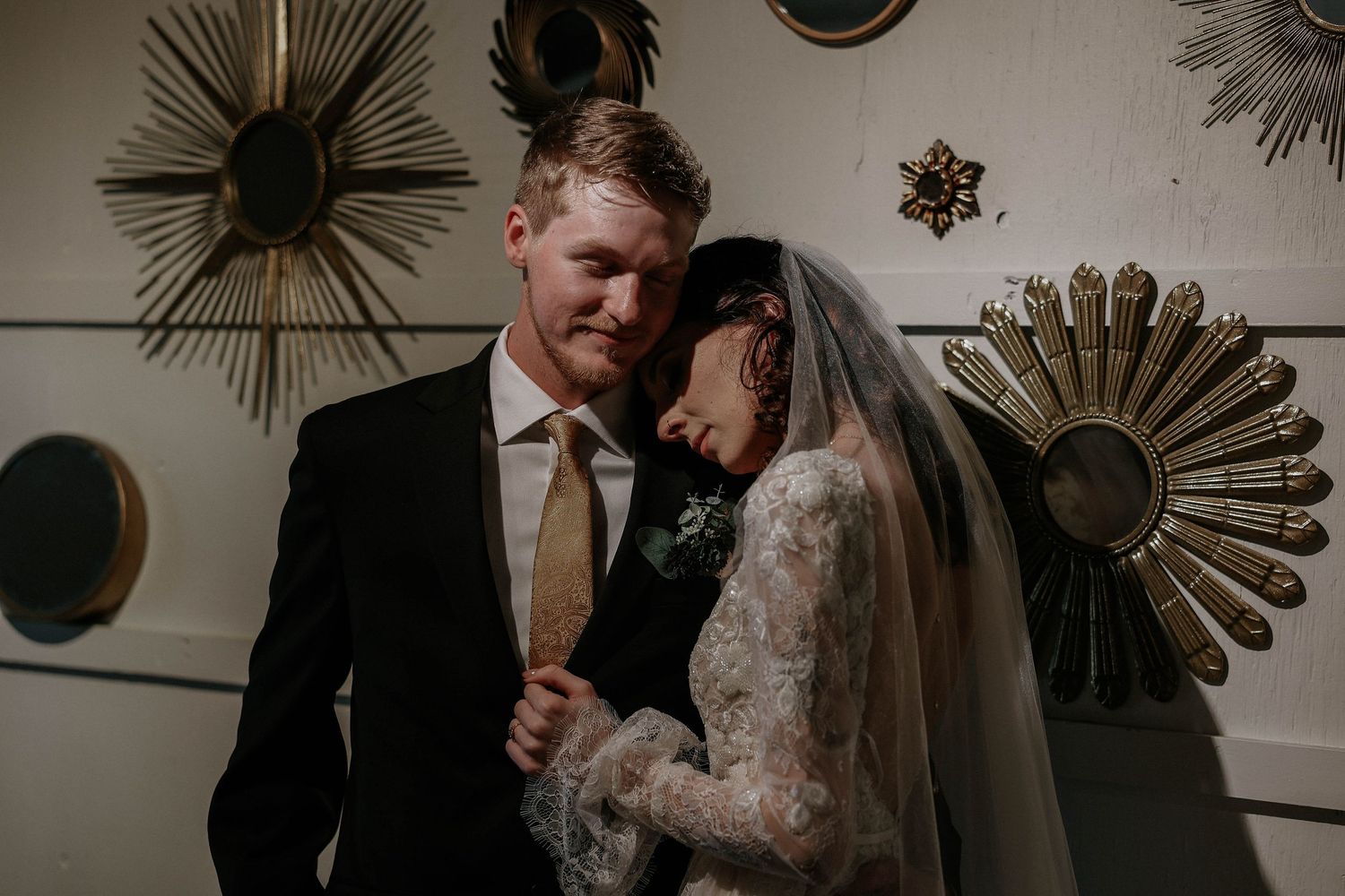 Bride and groom share a tender moment, bride in lace veil, groom in black suit with gold tie, sunburst mirrors on wall.
