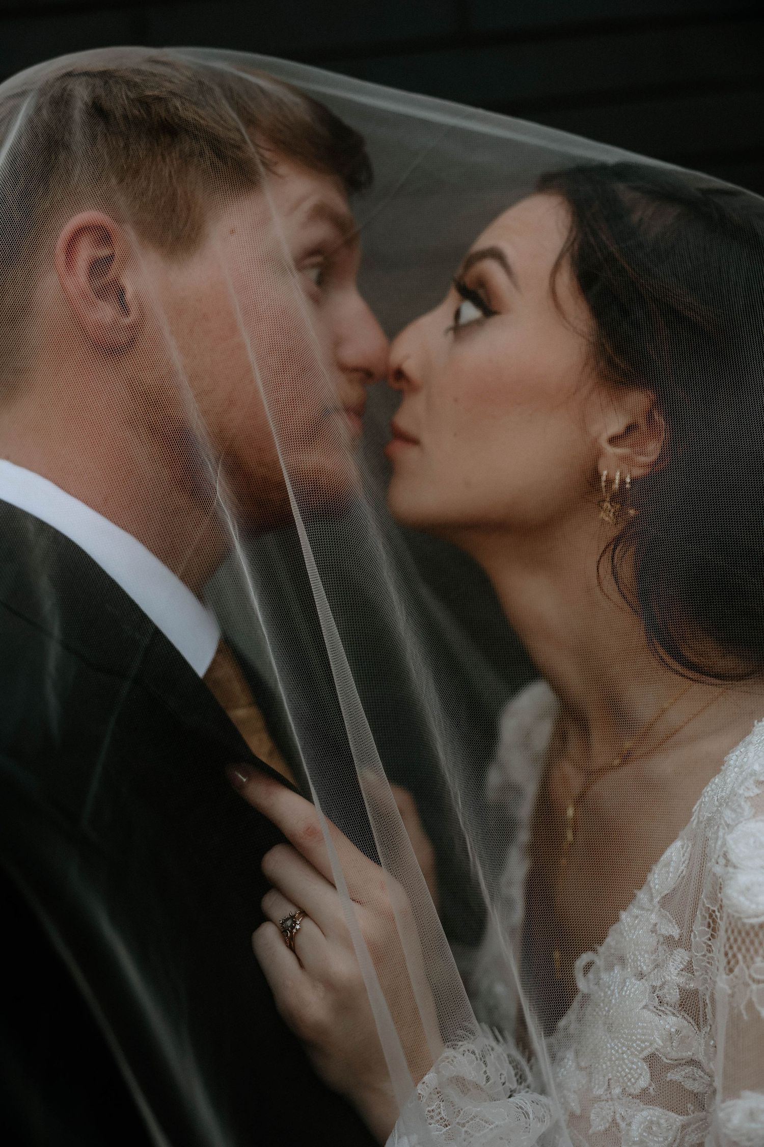 Bride and groom share a romantic almost-kiss under a sheer wedding veil in an intimate close-up portrait.
