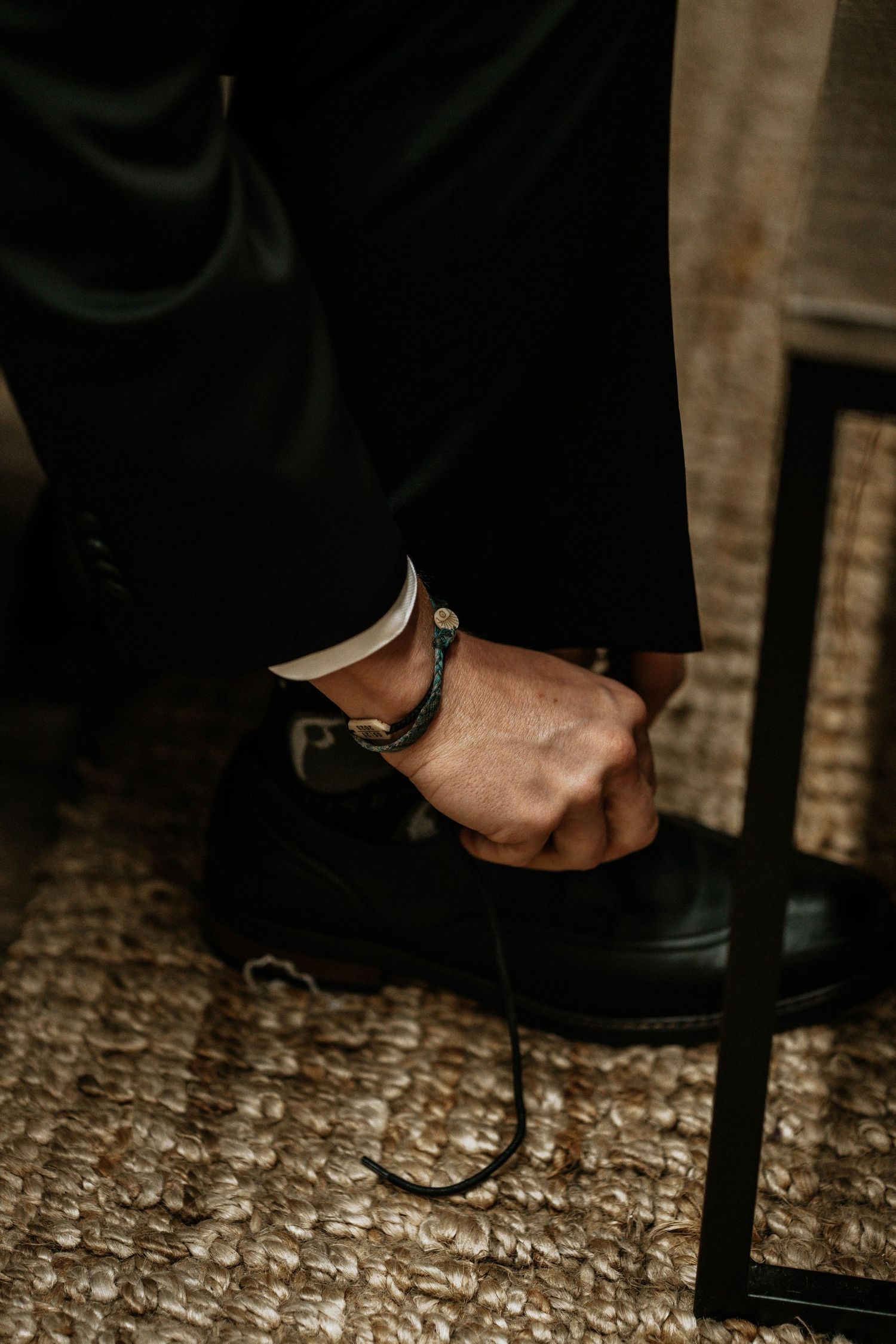 Groom tying black dress shoe laces wearing dark suit and leather bracelet on wrist.