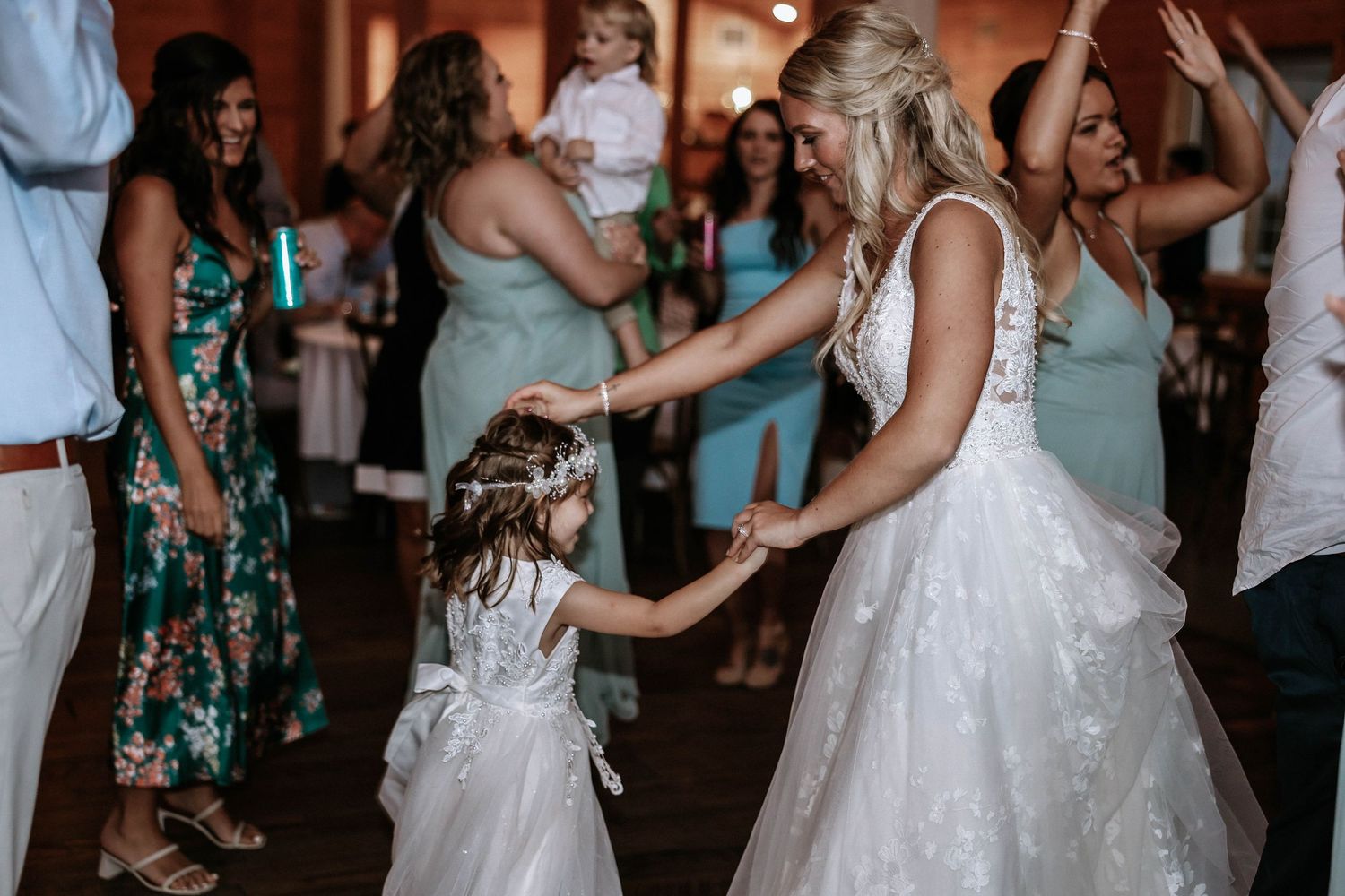 Bride in white gown dancing with flower girl at wedding reception while bridesmaids in sage green dresses look on.