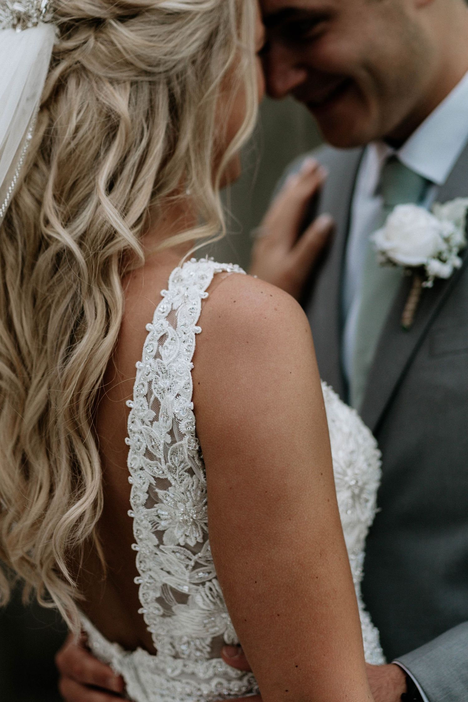 Bride in lace wedding gown with intricate strap detail embracing groom in gray suit outdoors.