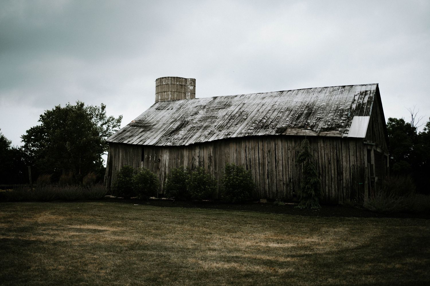 Rustic weathered wooden barn with metal roof and silo under overcast sky on rural farmland.