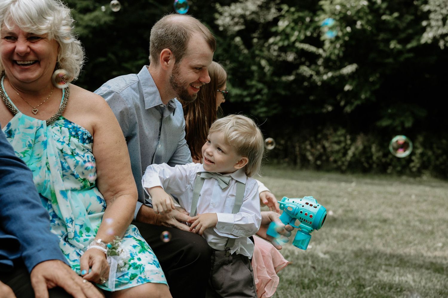 Family members smile as a toddler plays with a bubble gun at an outdoor wedding reception.