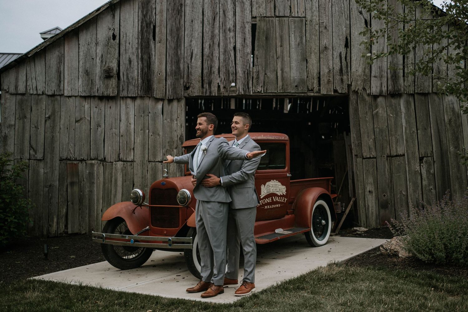 Two groomsmen in gray suits posing before a vintage red truck outside a rustic barn.